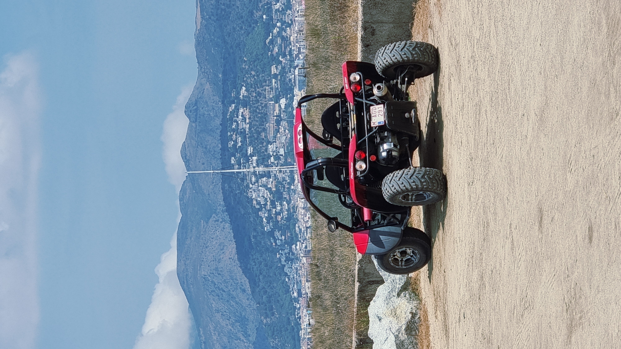 A red and grey buggy is parked on a dirt lot in front of a wall. The buggy is facing away from the camera, with its rear visible. It has large, knobby tires and a roll cage. Behind the buggy, a town with white buildings is spread across a hillside, with mountains in the background. A tall white mast, likely from a sailboat, rises above the town. The sky is partly cloudy with blue sky visible. The area appears to be outdoors, possibly in a coastal or hilly region.