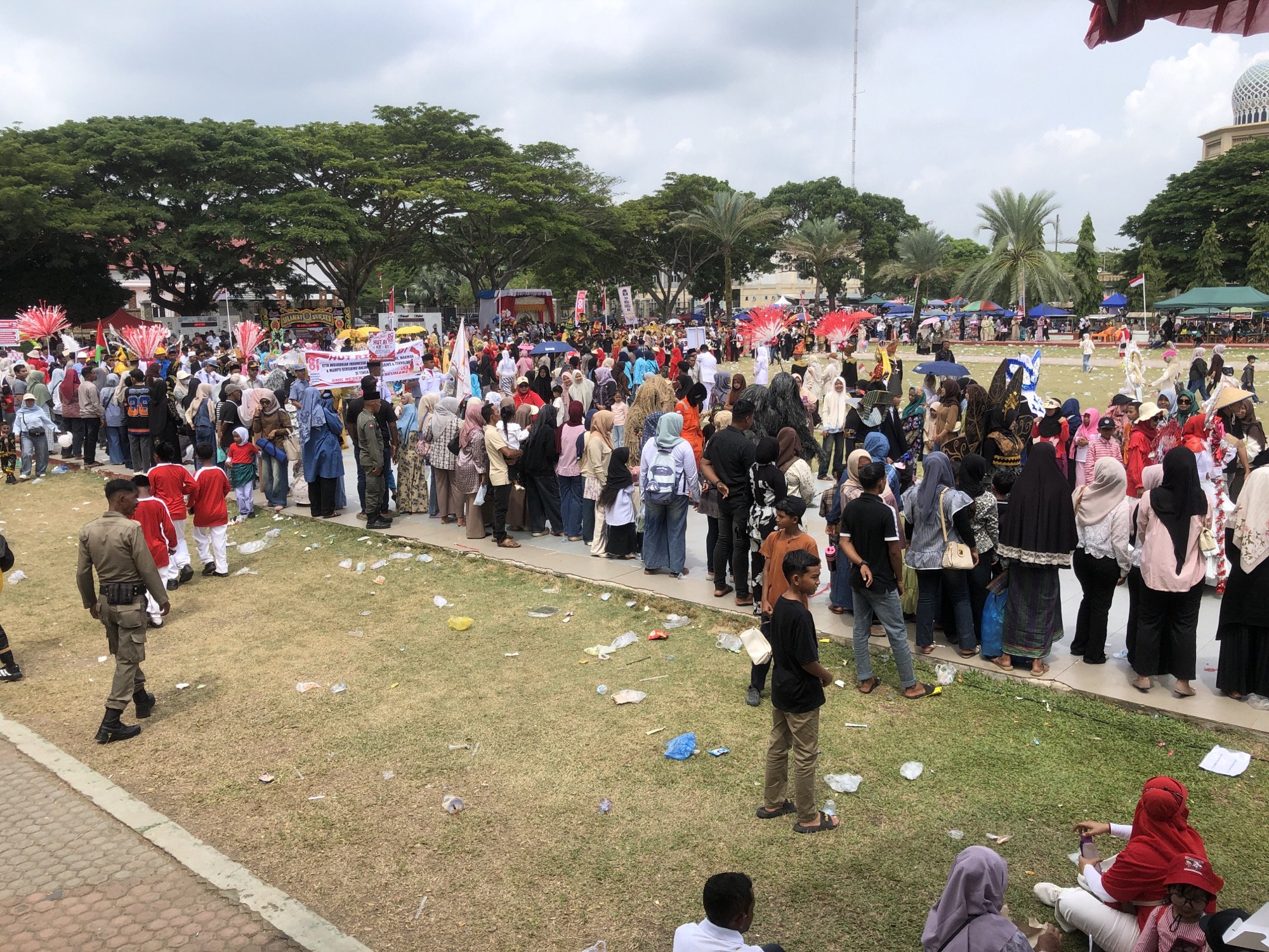 The image captures a daytime outdoor event with a large gathering of people on a grassy field in Lhokseumawe, Indonesia, likely near a mosque with a dome visible in the background. The crowd consists of a mix of men, women, and children, many wearing traditional Indonesian attire, including hijabs and sarongs. Some individuals are dressed in ceremonial costumes with elaborate headdresses and bright red feather decorations. The atmosphere appears festive, with banners and flags suggesting a parade or celebration. The ground is littered with various forms of trash, including plastic bags, cups, and paper debris, indicating a public event with significant consumption. In the foreground, a person in a military-style uniform is walking away from the camera, while others are standing and observing the event. The sky is overcast with scattered clouds.