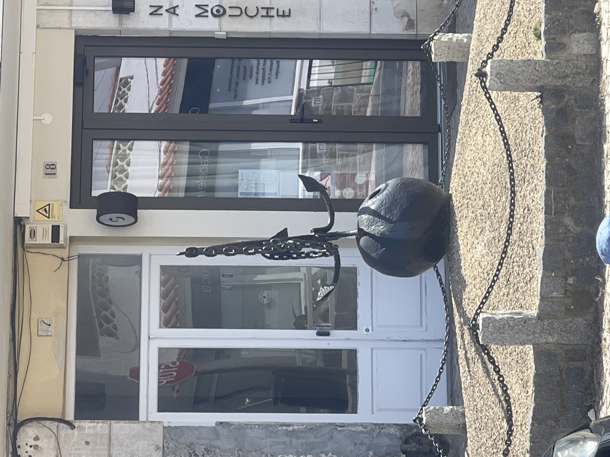 This image shows the exterior of a building with a street address of 18, located in Colares, Portugal. The building features a white double door on the left and a dark double glass door on the right. In front of the building, an anchor sculpture is displayed on a gravel bed, secured by chains and stone posts. A sign on the right side of the building reads "NA MOUCHE." The scene appears to be a commercial establishment, possibly a shop or restaurant, given the signage and display. The overall environment suggests a sunny day with shadows cast by the objects.