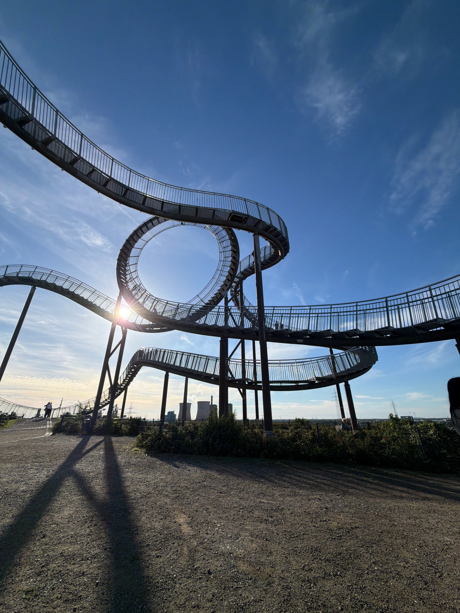 The image captures the Tiger & Turtle – Magic Mountain sculpture in the city of Duisburg, Germany, bathed in the warm light of a sunny day with a clear blue sky and scattered clouds. The sculpture, an artistic representation of a roller coaster, dominates the foreground and midground with its winding metal pathways and steep inclines. The sun's rays are visible, peeking through the central loop of the sculpture, creating a lens flare effect.
In the background, the industrial landscape of Duisburg is hinted at with the presence of cooling towers and power lines. The ground is covered in gravel, with a few sparse bushes and trees growing around the base of the sculpture. Two figures are visible on the pathway in the distance, walking away from the viewer, their shadows stretching long across the gravel. The overall scene evokes a sense of scale and architectural wonder against the backdrop of an industrial urban environment.