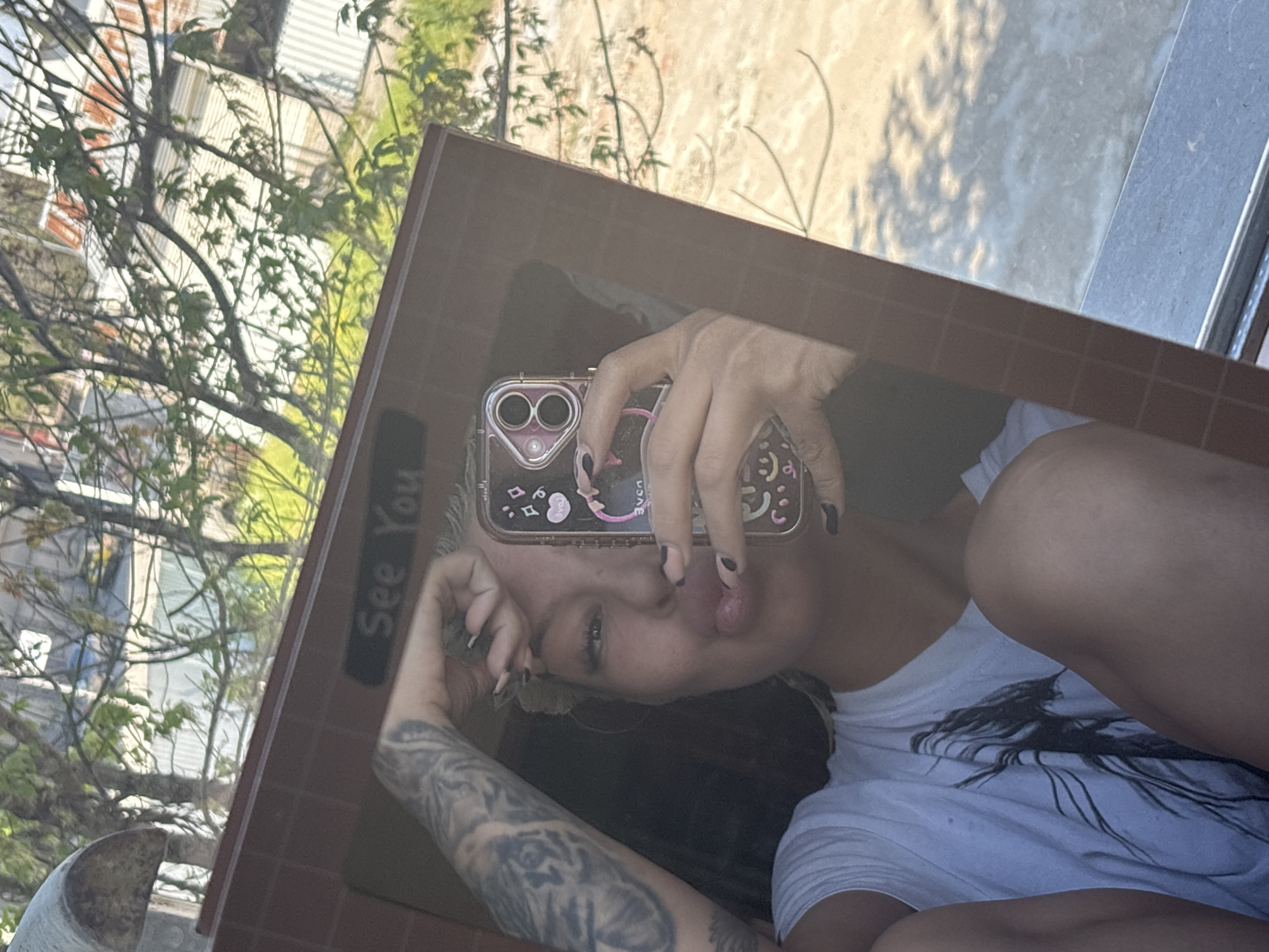 A woman is taking a selfie in a mirror. The mirror is framed in a red-brown border with the words "See You" written on it. The woman has blonde hair and is wearing a white t-shirt. Her left arm, visible in the mirror, is tattooed with a grey and black design. She is holding a phone with a decorated case in front of her face. The reflection shows that the woman is outside, possibly on a balcony. Trees and buildings are visible in the background, and what appear to be branches are in the upper left of the image. The location is possibly Plovdiv, Bulgaria, but there is no visual confirmation.