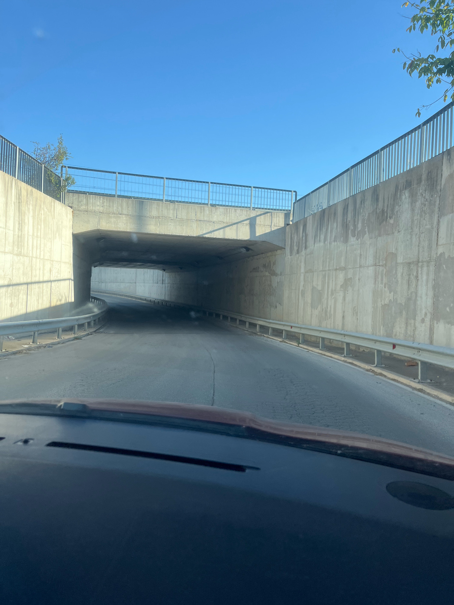 The image shows a view from inside a vehicle approaching the entrance of a tunnel. The tunnel is constructed with concrete walls and has a metal guardrail running along the side of the road. The road surface appears to be asphalt and shows some signs of wear and tear. The sky above is clear and blue, and some foliage from trees is visible on the right side of the frame, extending over the concrete structure. The scene is well-lit by natural sunlight, casting shadows on the tunnel walls. The interior of the vehicle is partially visible in the foreground, with the dashboard and windshield occupying the bottom portion of the image. There are no people or other vehicles visible in the immediate vicinity. The location is identified as being in the city of Sadovo, Bulgaria.