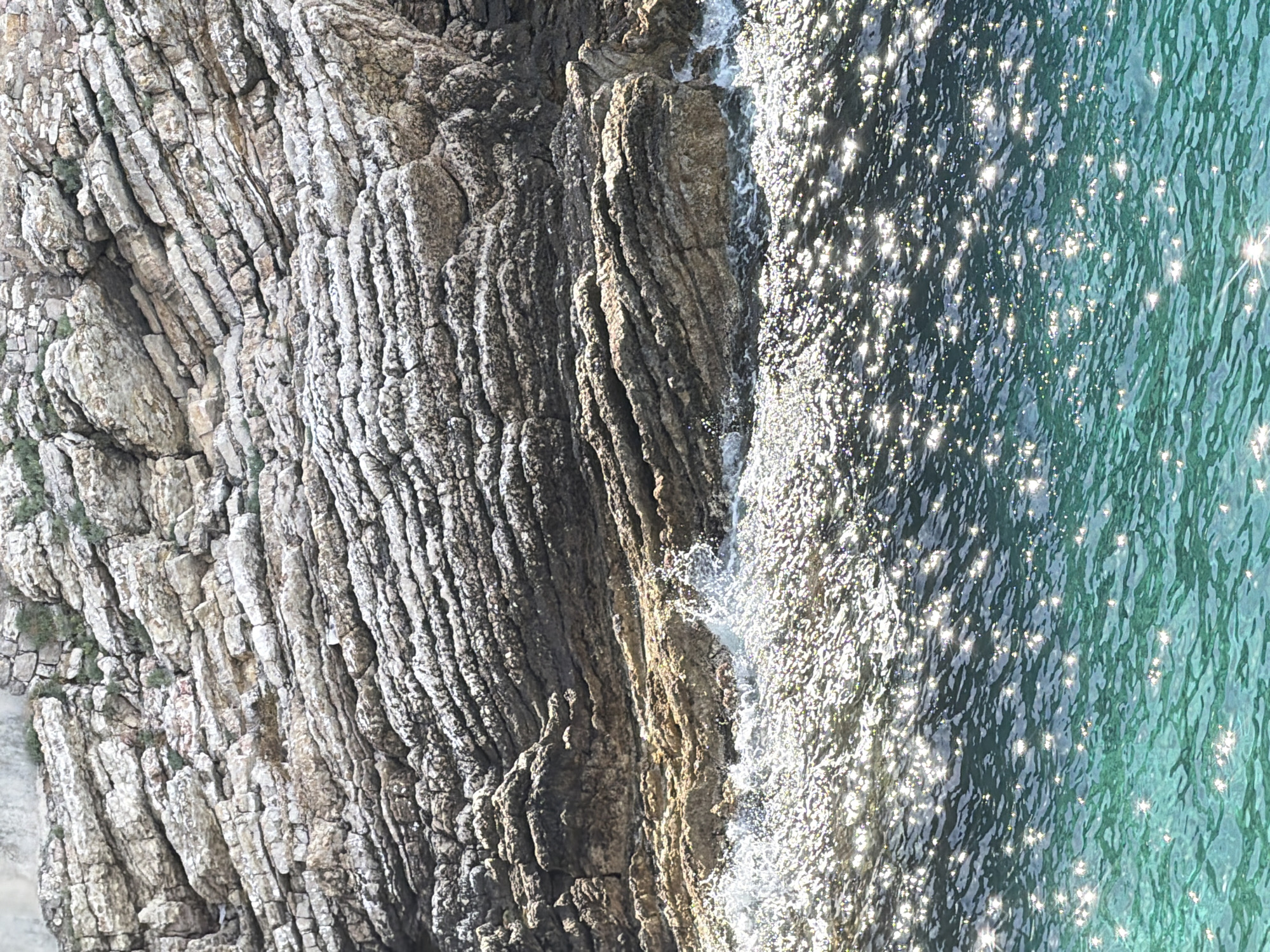The image shows a coastline in Petrovac centar, Montenegro. The left side of the frame is dominated by layered rock formations, transitioning from a lighter grey color on the far left to darker grey and brown nearer the water's edge. The right side of the frame contains water, the surface shimmering with reflected sunlight creating a sparkling effect. Small waves are visible where the water meets the rock. The rock formations are heavily textured with numerous small crevices and protrusions.