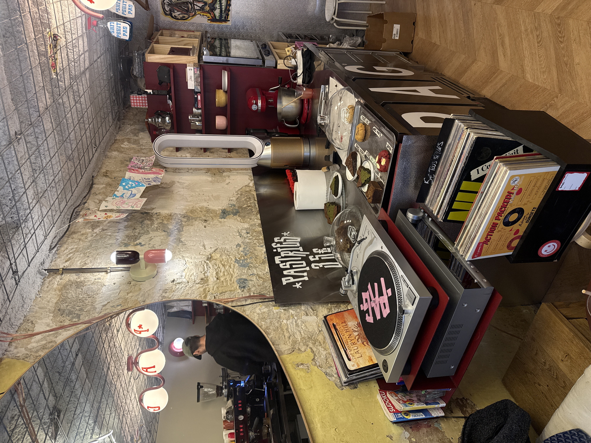 The interior of a small shop, possibly a pastry or coffee shop in Paris, France, is presented in an overhead perspective. The walls are stone and plaster, with exposed metal mesh reinforcement. Near the bottom of the frame, a mirror reflects a person in a black jacket, likely the barista, operating an espresso machine. A row of globe lights is visible on the wall above the mirror.
To the right of the mirror, a table is covered with vinyl records, a record player, and a sign reading "PASTRIES 3.5€". In the center of the scene, there's a tabletop arrangement that includes an air purifier, a pastry display, and other kitchen appliances. A red stand mixer and various shelves with dishes and kitchenware are visible in the background.
The floor is made of wood, and the overall ambiance of the space suggests a cozy and eclectic establishment with a focus on vinyl records and pastries. A sign with the letters "PAG" is prominently displayed on the front of the counter.