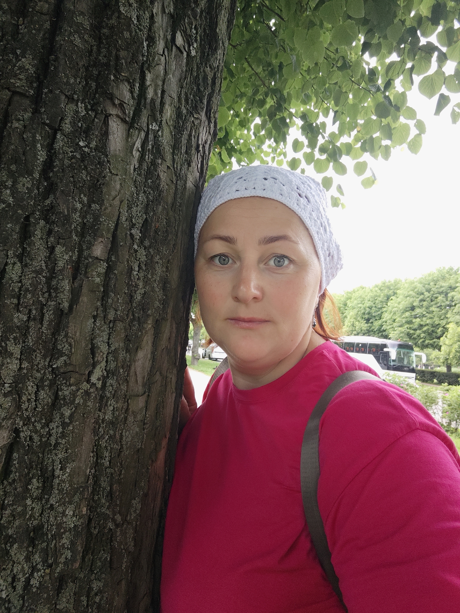A middle-aged woman with light skin and blue eyes is shown in a close-up portrait. She is wearing a white crocheted hat and a bright pink t-shirt. She is standing next to a large tree with rough bark. The background is slightly out of focus, showing green foliage from trees overhead and a hazy view of a street with a bus and a car. The sky is overcast. The image appears to be taken outdoors in a park or on a tree-lined street. There is no specific indication of the location being Saint Petersburg, Russia.