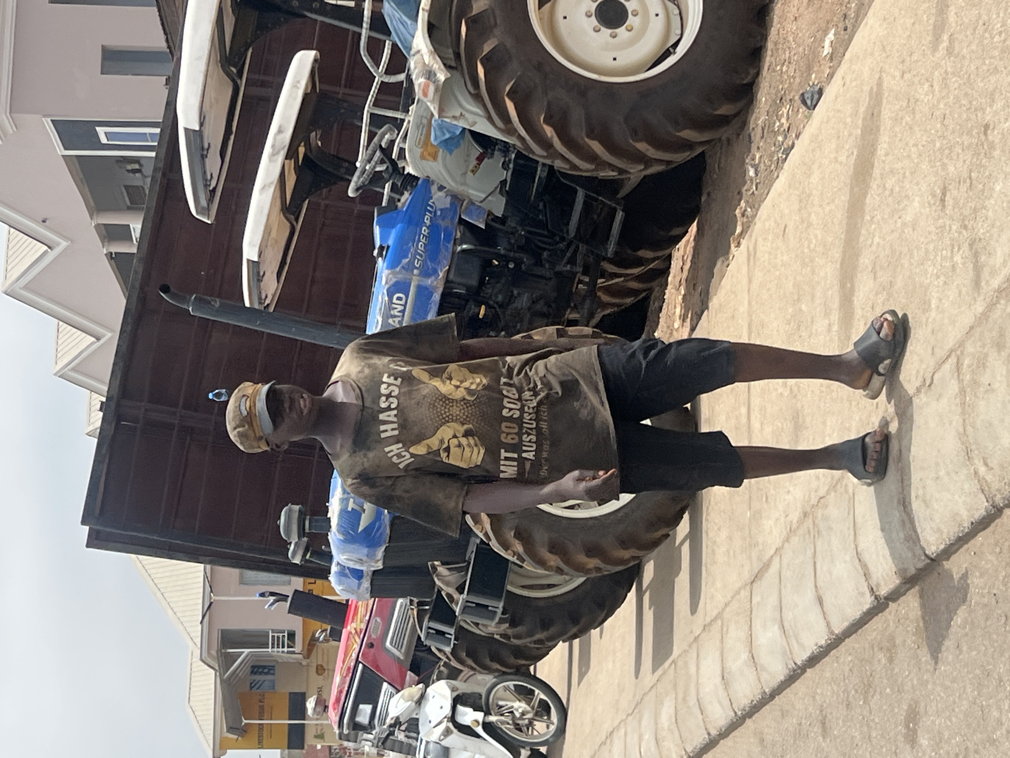 A man is standing on a sidewalk in the city of Jos, Nigeria, in front of a row of parked tractors. He is wearing a t-shirt with an image of two hands pointing toward the text "ICH HASSE MIT 60 SOLL AUSZUSEEN" and black shorts, along with sandals. The tractors are various colors, including red, blue, and white. The background includes buildings of mixed construction. The overall tone is a candid snapshot of daily life.