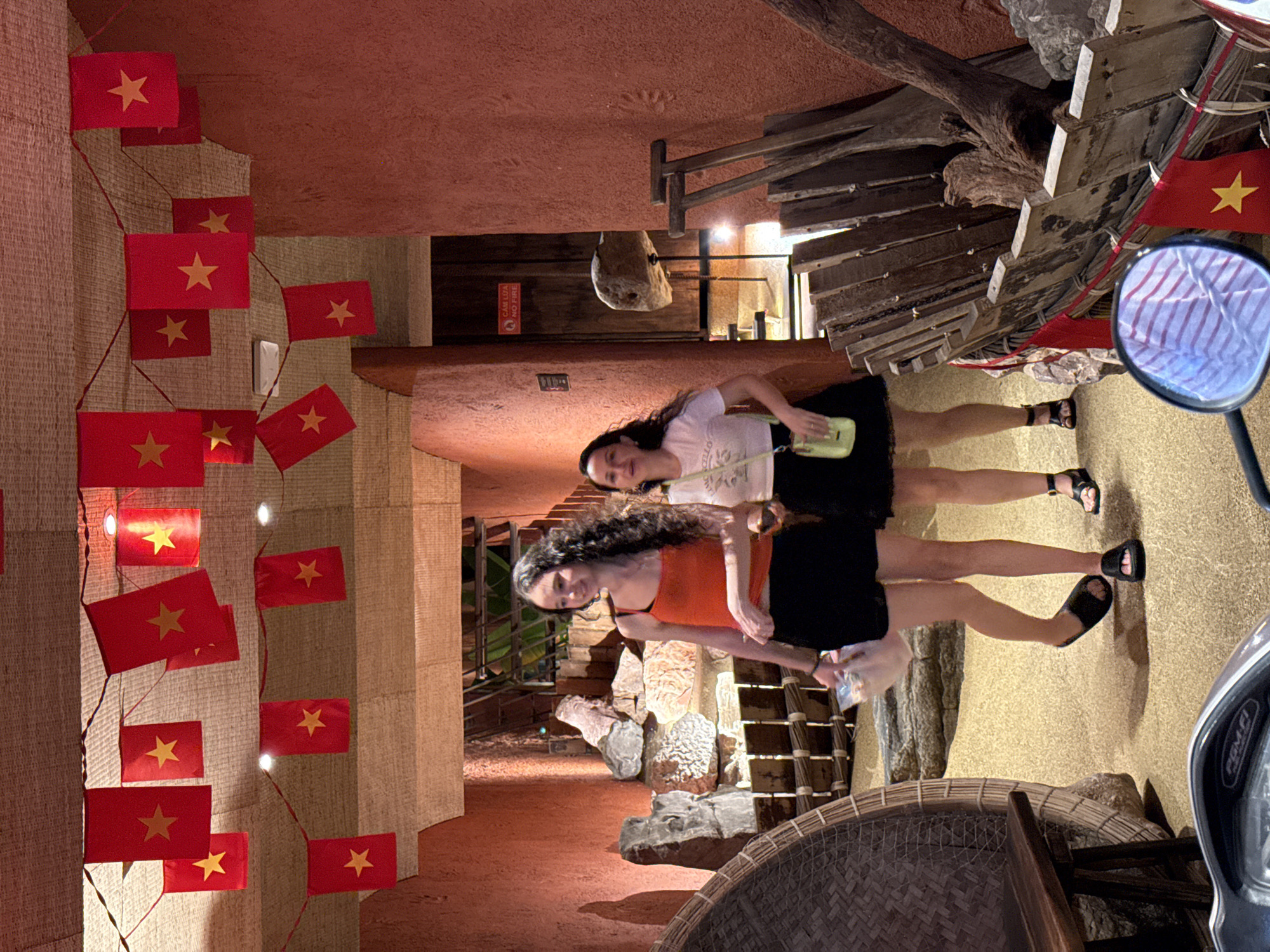 Two women stand in an indoor space decorated with Vietnamese flags, with the red flags featuring a gold star hung along a wall. The space features exposed wooden beams and what appears to be a natural stone or rocky formation along the interior design. One woman is wearing an orange tank top and a black skirt, while the other is wearing a white shirt and a black skirt, with a small green bag slung across her shoulder. A basket-like object is in the foreground. The location is Nha Trang Ward, Vietnam.