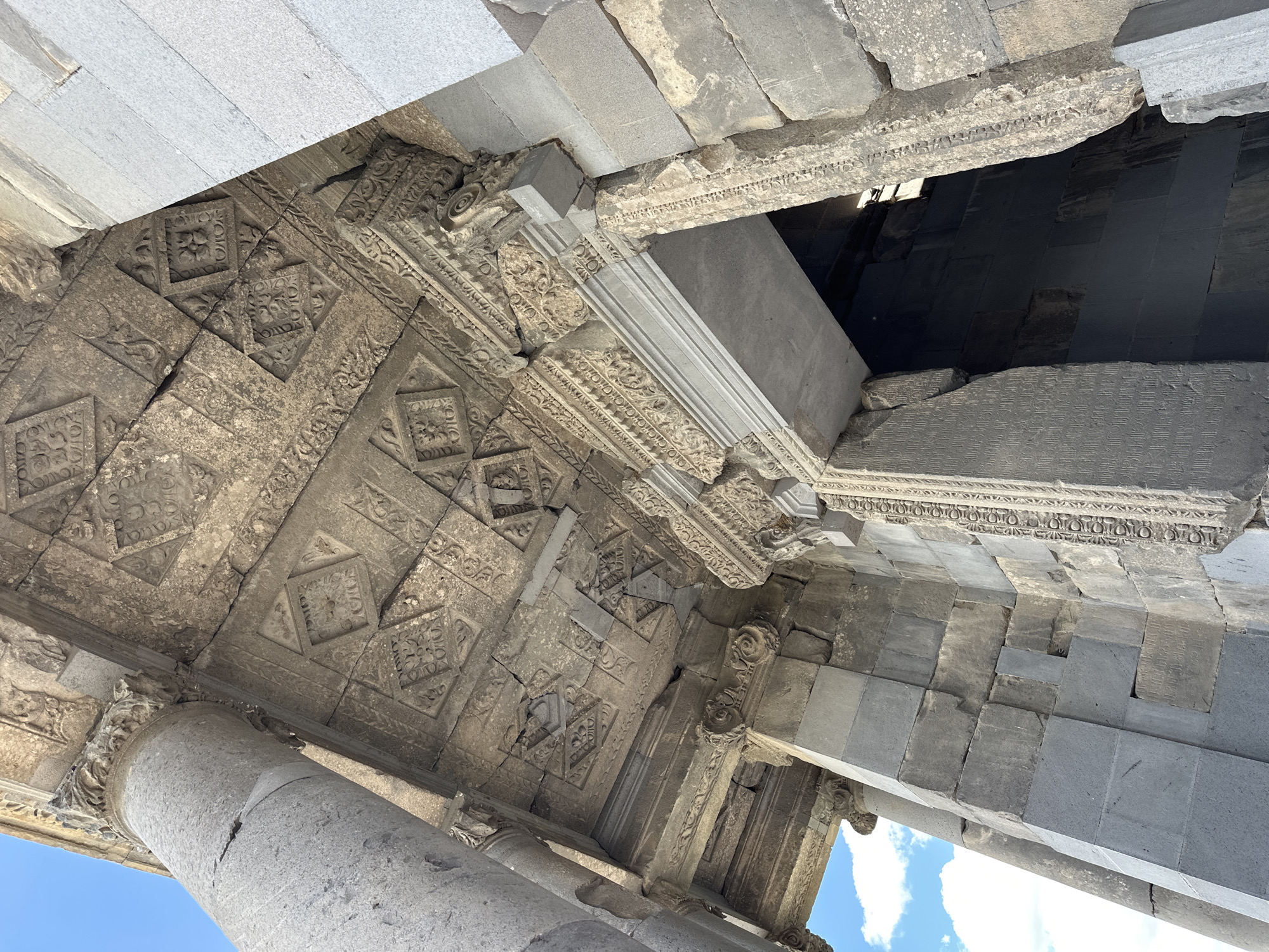 The image shows a close-up, low-angle view of the entrance to the Temple of Garni in Garni, Armenia. The temple, constructed of grey basalt blocks, features intricate stone carvings on its facade and ceiling. The ceiling is adorned with geometric patterns and floral motifs carved into individual stone panels. A large, fluted column stands to the left of the entrance, partially obscuring the sky, which is blue with scattered white clouds. The entrance itself is a large, arched doorway framed by carved stone. The weathered stone suggests the age of the structure. The light suggests it is daytime.