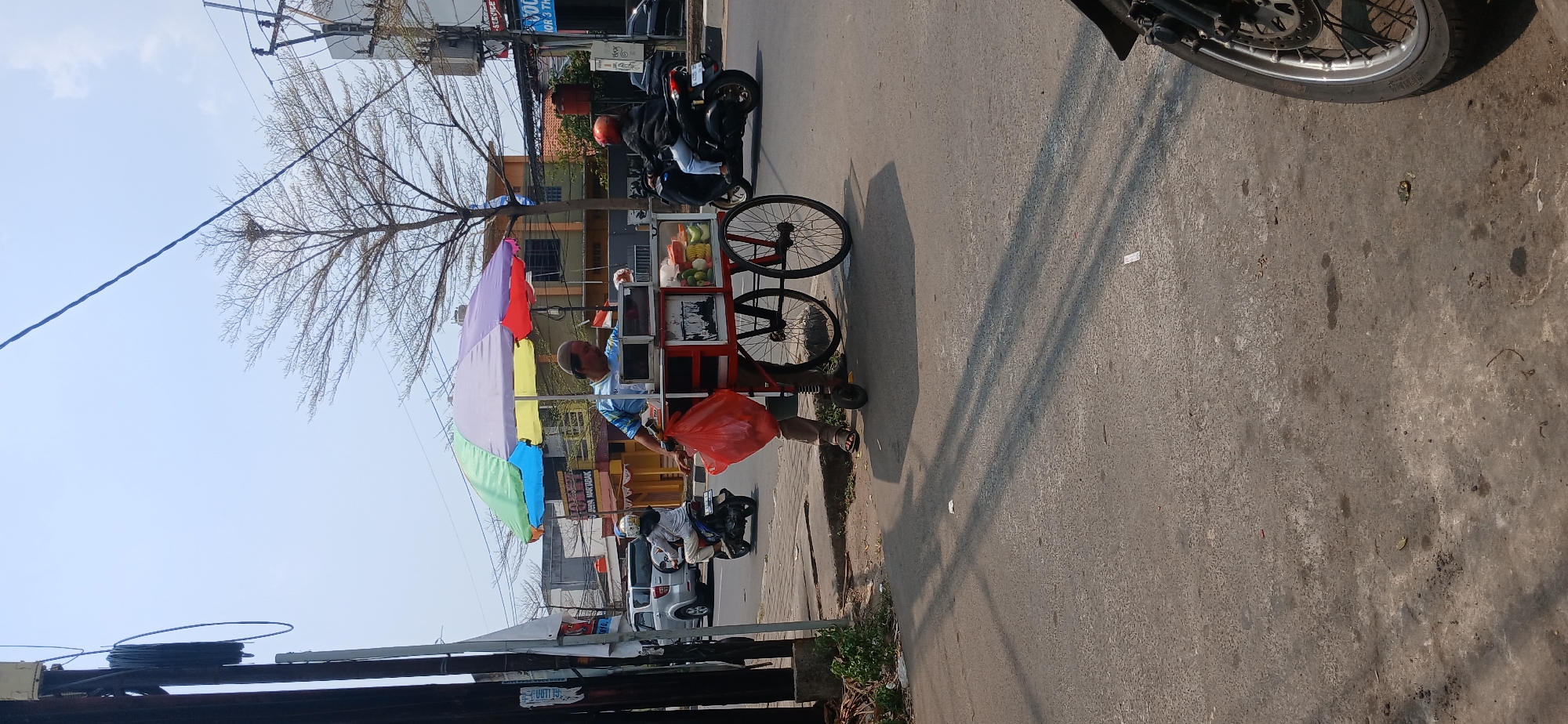 In Baros, Indonesia, a man stands beside a colorful, multi-umbrella-covered cart selling what appears to be fruit. He holds a red plastic bag in his left hand and wears a light blue and white striped shirt, brown pants, and a beige cap. The cart has a white top with compartments, displaying various fruits, including what look like bananas and green limes, and red apples. Behind him, a silver SUV is parked on the side of the road. A woman on a black scooter passes by in the street, wearing a helmet and a light gray jacket. The scene takes place on a street with a paved road and sidewalk, under a bright, clear sky. Overhead, power lines and a bare-branched tree are visible. The overall setting suggests a sunny day in a bustling urban environment.