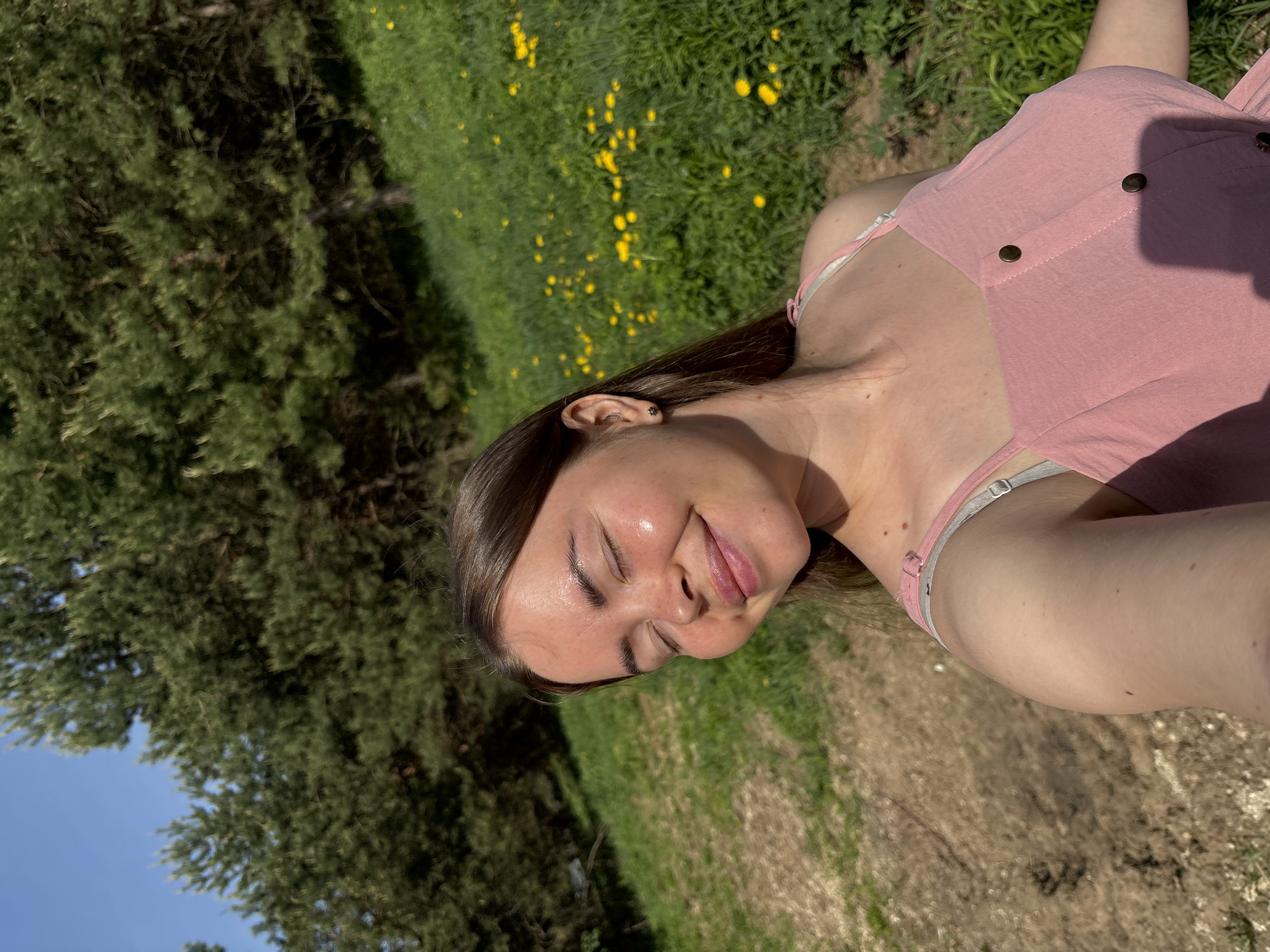 A young woman is lying in a grassy area with yellow flowers near trees. She is wearing a pink dress and has her eyes closed. The photo appears to be taken outside in the daylight. The location in the city of Русский Вожой, Russia, is not evident from the image.