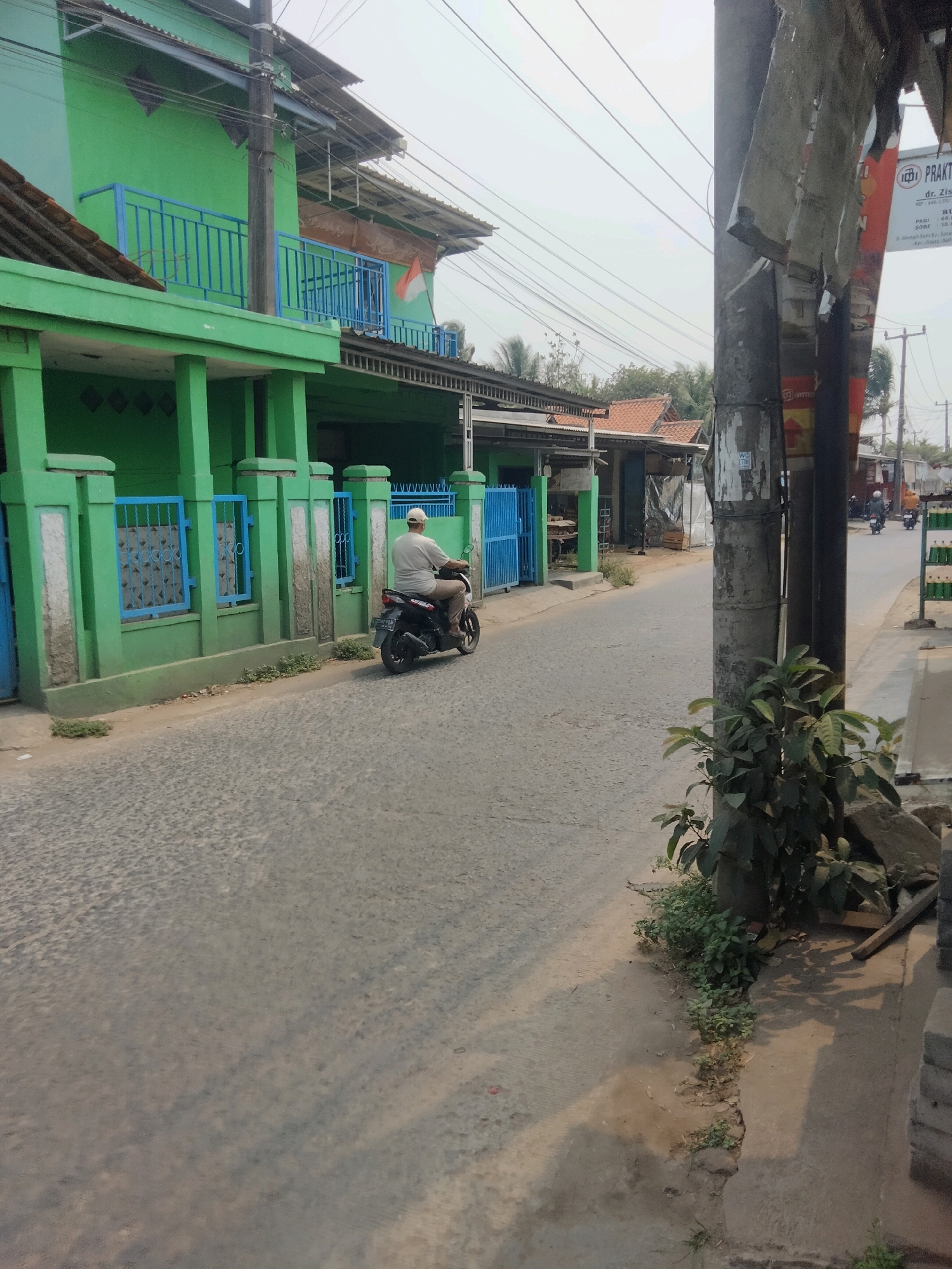 A side view shows a person riding a black motorcycle down a paved road in Suka Sari, Indonesia. The rider, wearing a light-colored t-shirt and a cap, is facing away from the camera. A vibrant green building with blue railings lines the left side of the road, featuring a concrete fence with blue bars. The building appears to be multi-story and has a tiled roof. On the right side of the road, a utility pole is prominent, with a faded poster attached to it. Further down the road, other buildings and motorcycles are visible. The sky is overcast, suggesting a cloudy day. The ground is uneven, with some vegetation growing along the roadside.