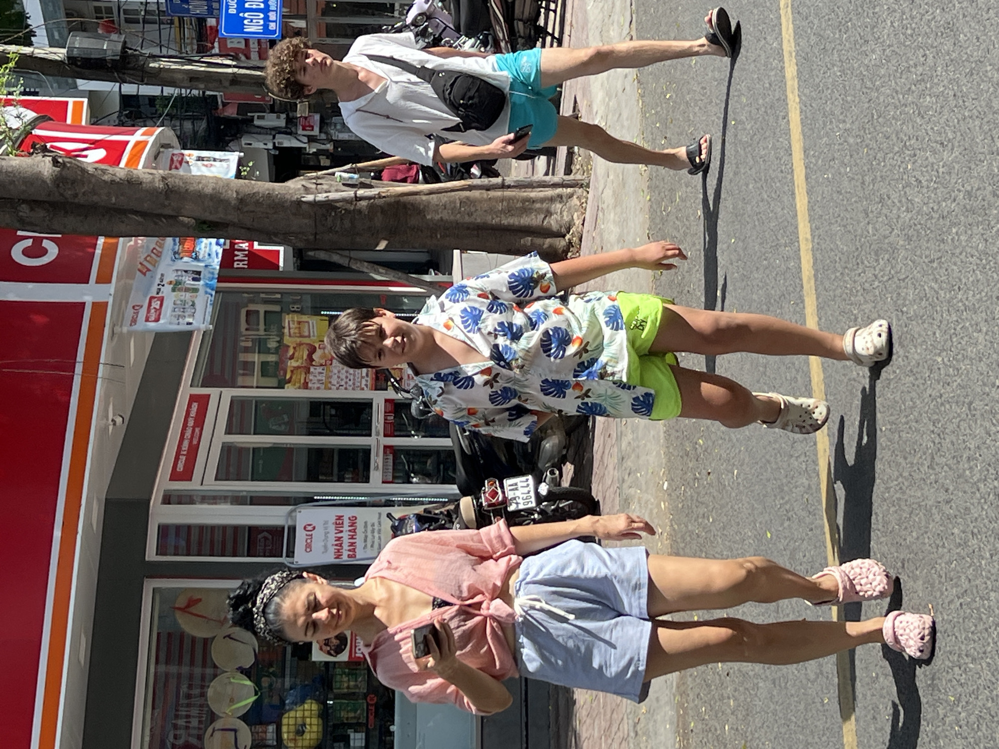 Three individuals are walking on a paved street in Nha Trang, Vietnam, with a store in the background. The store has a red awning and signage. The person on the left, a woman with dark hair, is wearing a pink button-down shirt, light blue shorts, and pink clogs. She is holding a smartphone. In the center, a young person with brown hair is wearing a short-sleeved floral shirt and neon green shorts, with beige clogs. To the right, a young man with curly brown hair is wearing a white hooded shirt, turquoise shorts, and black sandals, with a black sling bag across his chest. Motorcycles are parked in front of the store. A tree trunk is visible between the store and the street. A yellow line marks the edge of the road on the asphalt.