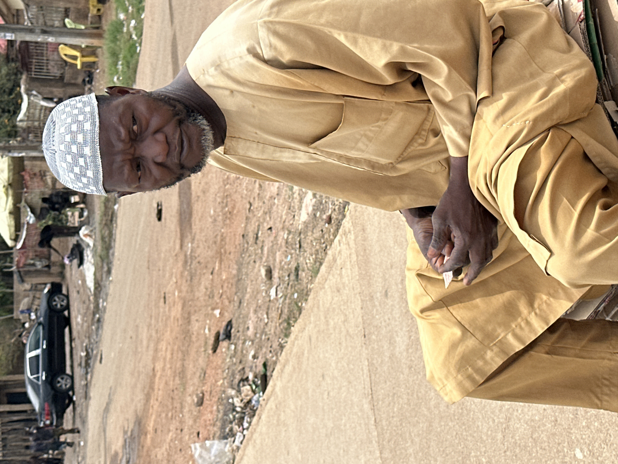 An outdoor, daytime portrait captures an older Black man, likely in Jos, Nigeria, looking directly at the camera with a serious and slightly furrowed expression. He wears a light tan or beige traditional tunic, possibly a boubou or kaftan, and a white and light blue patterned kufi cap. A short grey beard frames his face. His dark-skinned hands are clasped in his lap, holding a small, unidentifiable piece of white paper or fabric.
The man appears to be seated on or next to some cardboard boxes on the right side of the frame. The setting is an unpaved, dusty road on the left, contrasting with a lighter, possibly concrete or paved strip in the foreground where the man is situated. The background reveals hints of sparse vegetation and scattered debris or trash. On the far left, a black sedan is partially visible, parked on the dirt road. The clear lighting indicates it is daytime. The overall scene suggests a roadside or semi-urban environment. No readable text is visible in the image.