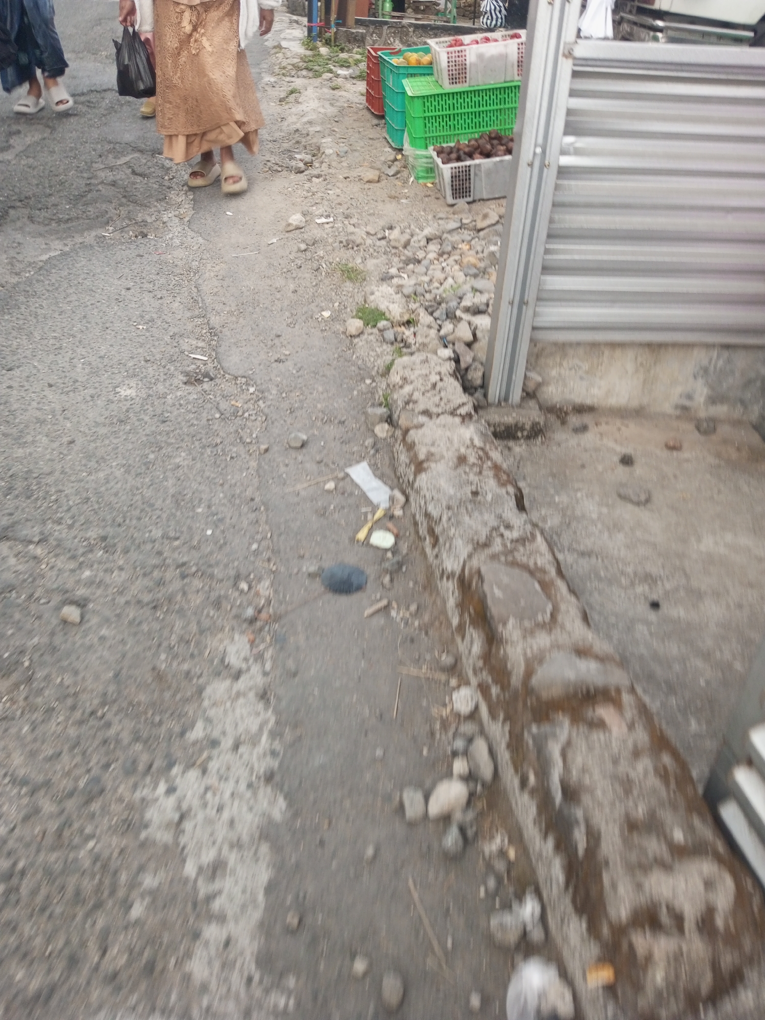 The image depicts an outdoor scene on a street in Pandansari, Indonesia. The ground is primarily asphalt, with sections of rougher, pebbly material. In the foreground, a concrete curb runs along the edge of the street, appearing weathered and partially covered in dirt and debris. Scattered across the ground are various small stones, bits of trash, and a yellow object that may be a piece of fruit.
In the midground, a person wearing a tan-colored dress and sandals is walking away from the camera, carrying a black plastic bag. Another person, only their legs are visible, wearing a dark blue garment and white sandals, is walking to the left in the background.
Further back, stacked green and red plastic crates are visible, some containing what appear to be fruits like rambutans and possibly oranges. These crates are placed on the ground near a corrugated metal barrier, suggesting a market or street vendor setting. The overall impression is a glimpse of everyday life on a street in Pandansari, likely near a commercial area.