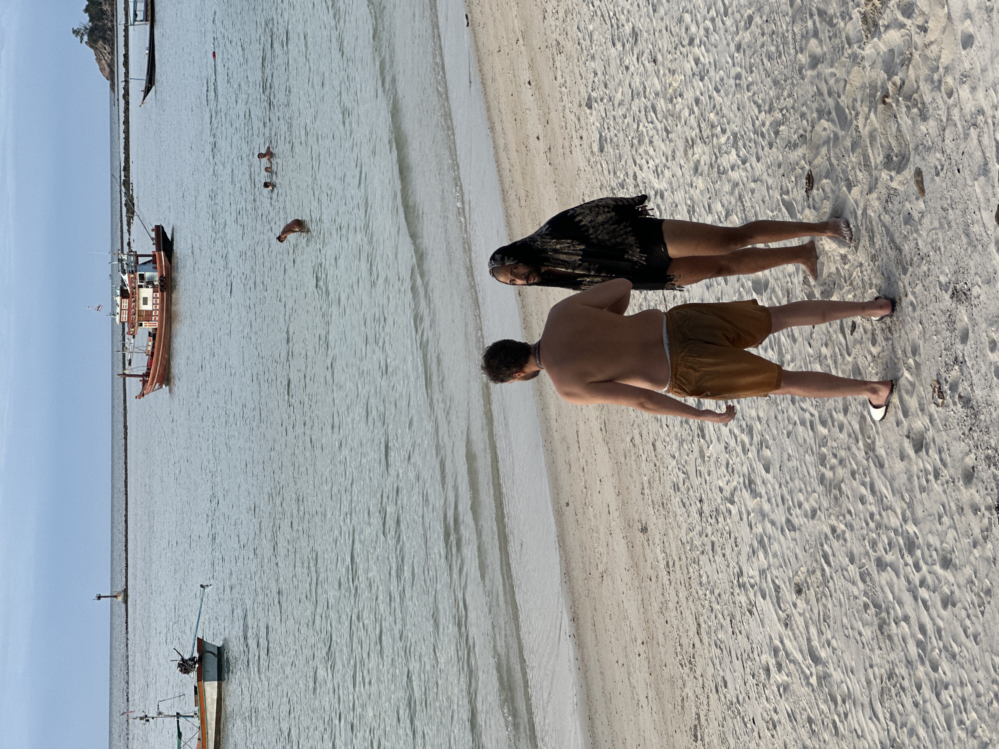 Two individuals are standing on a sandy beach, facing each other. The person on the left has their back to the camera, wearing tan shorts and white flip-flops. The person on the right is facing the camera, wearing black shorts and a patterned black wrap around their head and shoulders. The beach is a light tan color with visible footprints. In the background, the ocean is calm with small waves rolling onto the shore. Several boats are visible in the water, including a large red and white fishing boat in the middle distance and smaller boats closer to the shore. A lighthouse-like structure and a rocky outcrop with vegetation are visible on the horizon. The sky is overcast. The scene is likely located in ตำบลเกาะพะงัน, Thailand, given the presence of traditional Thai fishing boats and the tropical beach environment.