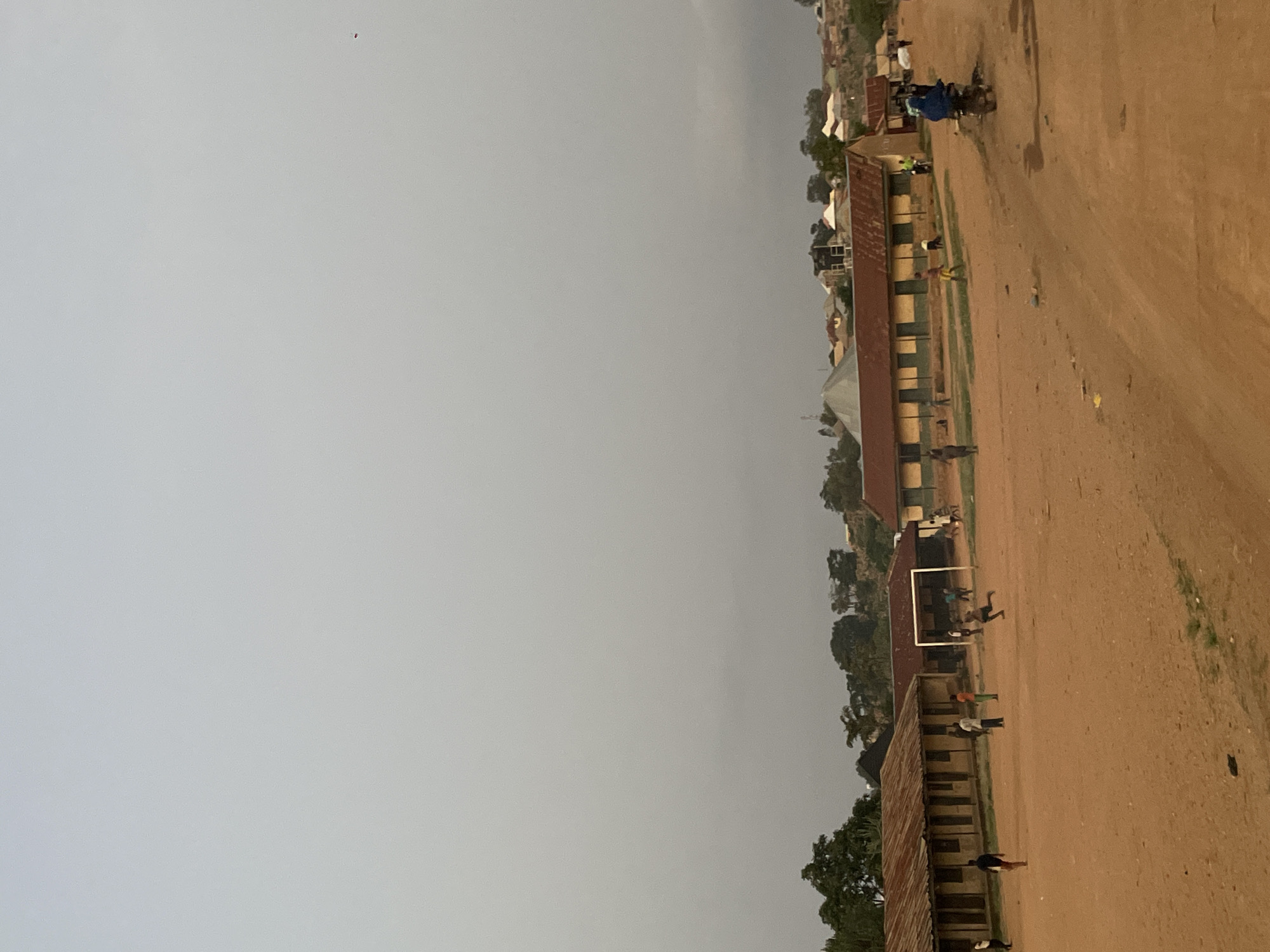 The image shows a street scene in Barikin Gidar Kanat, Nigeria. A dirt road runs along the right side of the frame. Buildings, possibly schools or residential structures, line the other side. People are scattered along the road and near the buildings. A soccer goal is visible between two buildings. The sky is overcast. A motorcycle is on the right edge of the frame.