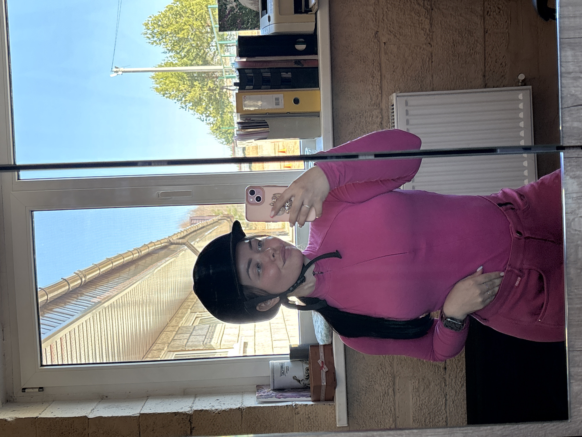 A young woman takes a mirror selfie indoors in Русский Вожой, Russia, during what appears to be a bright, sunny day. She is wearing a bright pink long-sleeved top or sweatshirt and matching pink pants, along with a black equestrian helmet, under which her dark hair is pulled back into a ponytail. She holds a rose gold smartphone, equipped with a decorative case and a ring holder, in her right hand to capture the reflection. A silver ring is visible on her left ring finger, and a smartwatch is on her left wrist.
The reflection reveals a window behind her, offering a view of a clear blue sky, vibrant green tree foliage, and a utility pole with wires. Below the outdoor scene, a house roof with light-colored vertical siding and brown gutters is visible through the lower part of the window. Inside, a bookshelf filled with various binders and files, including yellow and black ones, is reflected above her head. A white radiator is mounted on a textured brown wall behind her. On the windowsill, a white container with the partial text "TIMACY" and a small brown gift box are visible. The woman has a calm expression, with a slight smile. A small red tag, possibly with text like "RED GOOSE," is on the pocket of her pink top.