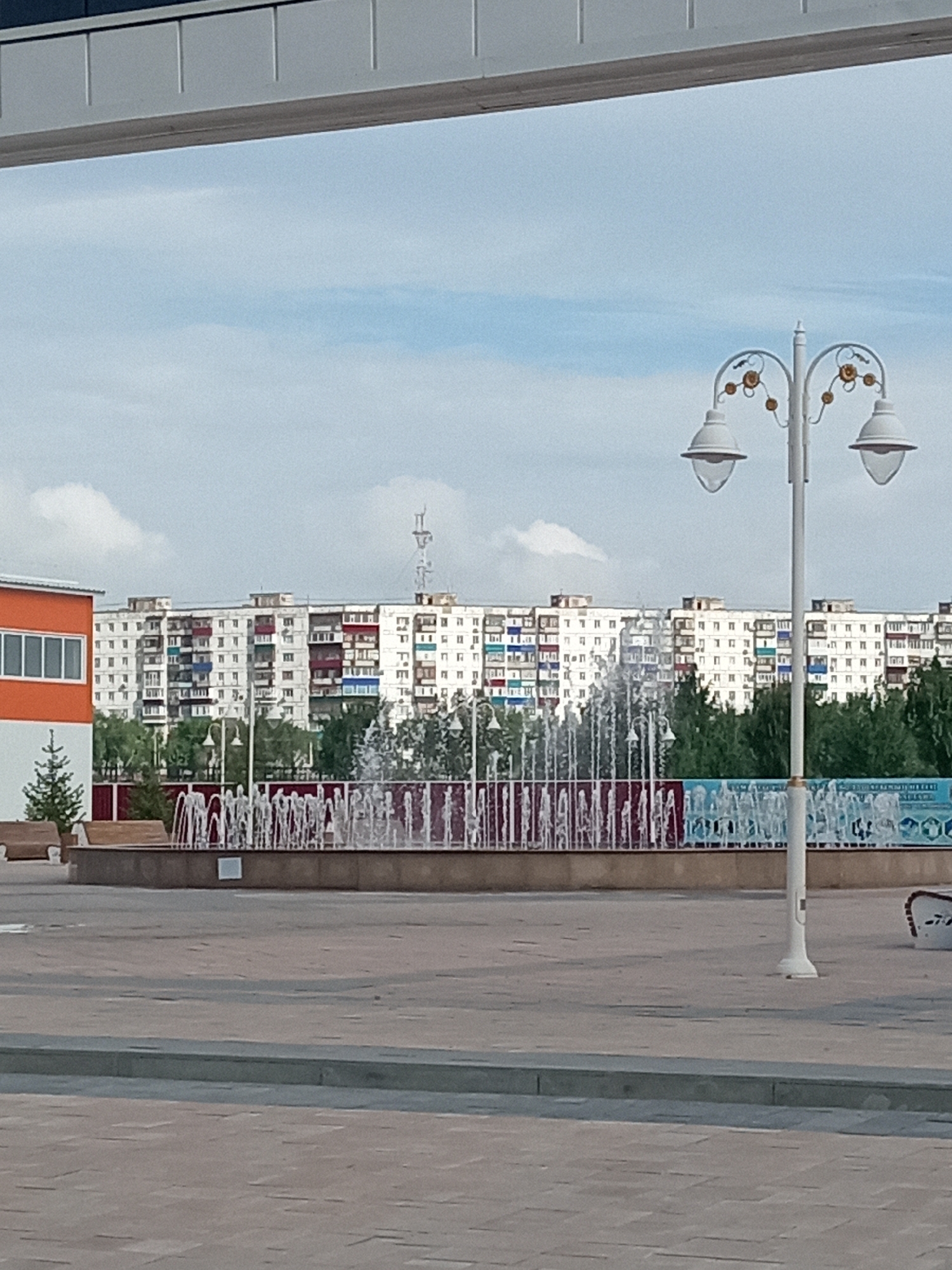 The image depicts an outdoor urban scene in Rudny, Kazakhstan, featuring a multi-story apartment building in the background and a decorative fountain in the mid-ground. The apartment building is predominantly white with balconies and windows that have colorful accents, suggesting a Soviet-era architectural style. In front of the apartment building, a line of green trees is visible. The fountain is active, with jets of water spraying upwards. A lamppost with two lights and decorative elements stands to the right of the fountain. In the foreground, a paved walkway or plaza is visible, with a low ledge. To the left, a portion of an orange and white building is seen. The sky is partly cloudy. There are no people visible in the image.