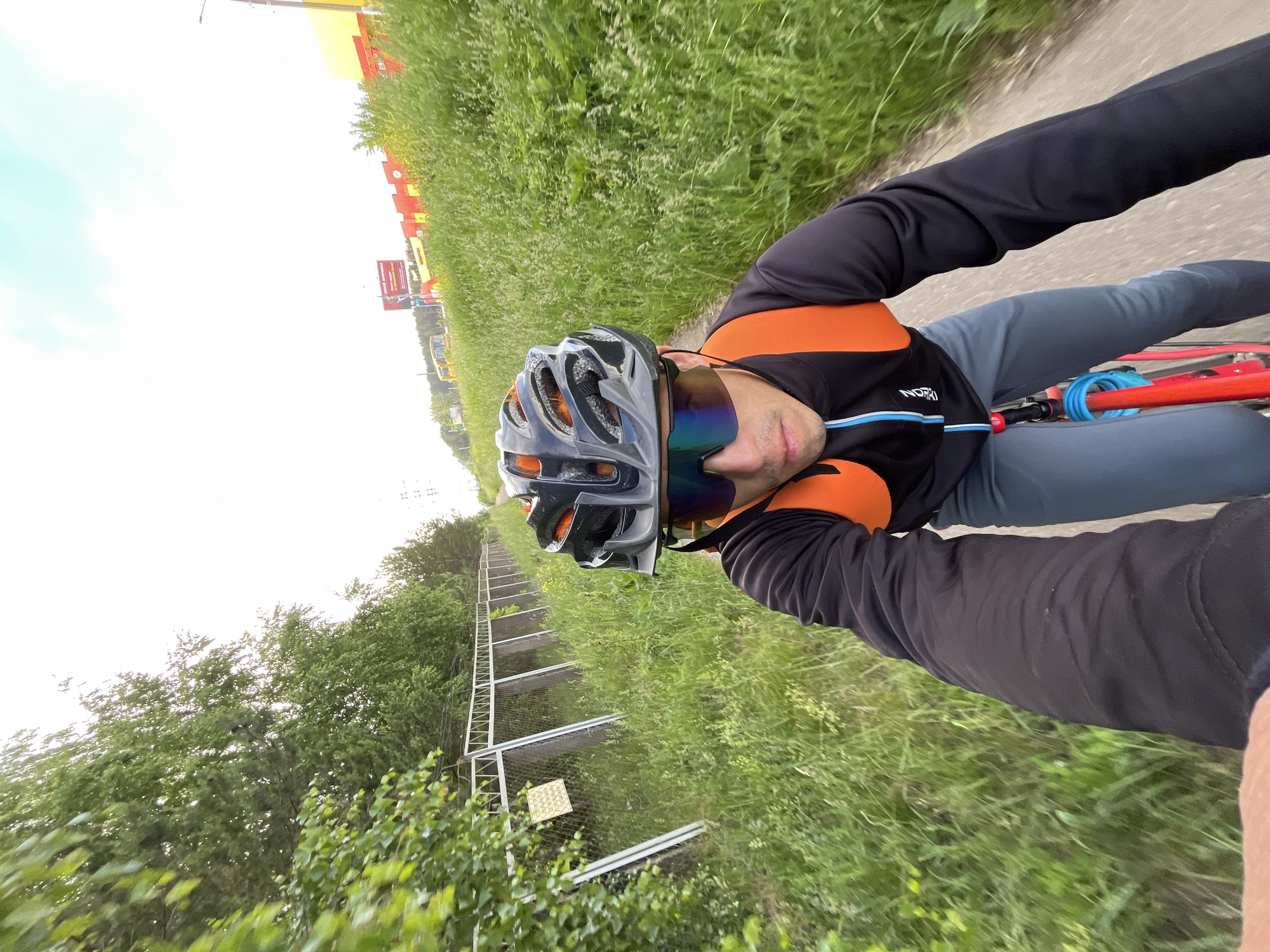 A male cyclist wearing a black jacket, grey pants, a black and orange helmet, and mirrored sunglasses is riding a red bicycle on a dirt path next to a grassy area. The cyclist is wearing an orange backpack. The path is bordered by a metal fence on the left and tall grass and trees on the right. In the background, there are buildings with red and yellow accents, a billboard, and some trees. The sky is overcast. The location is identified as the city of Chaikovsky, Russia.
