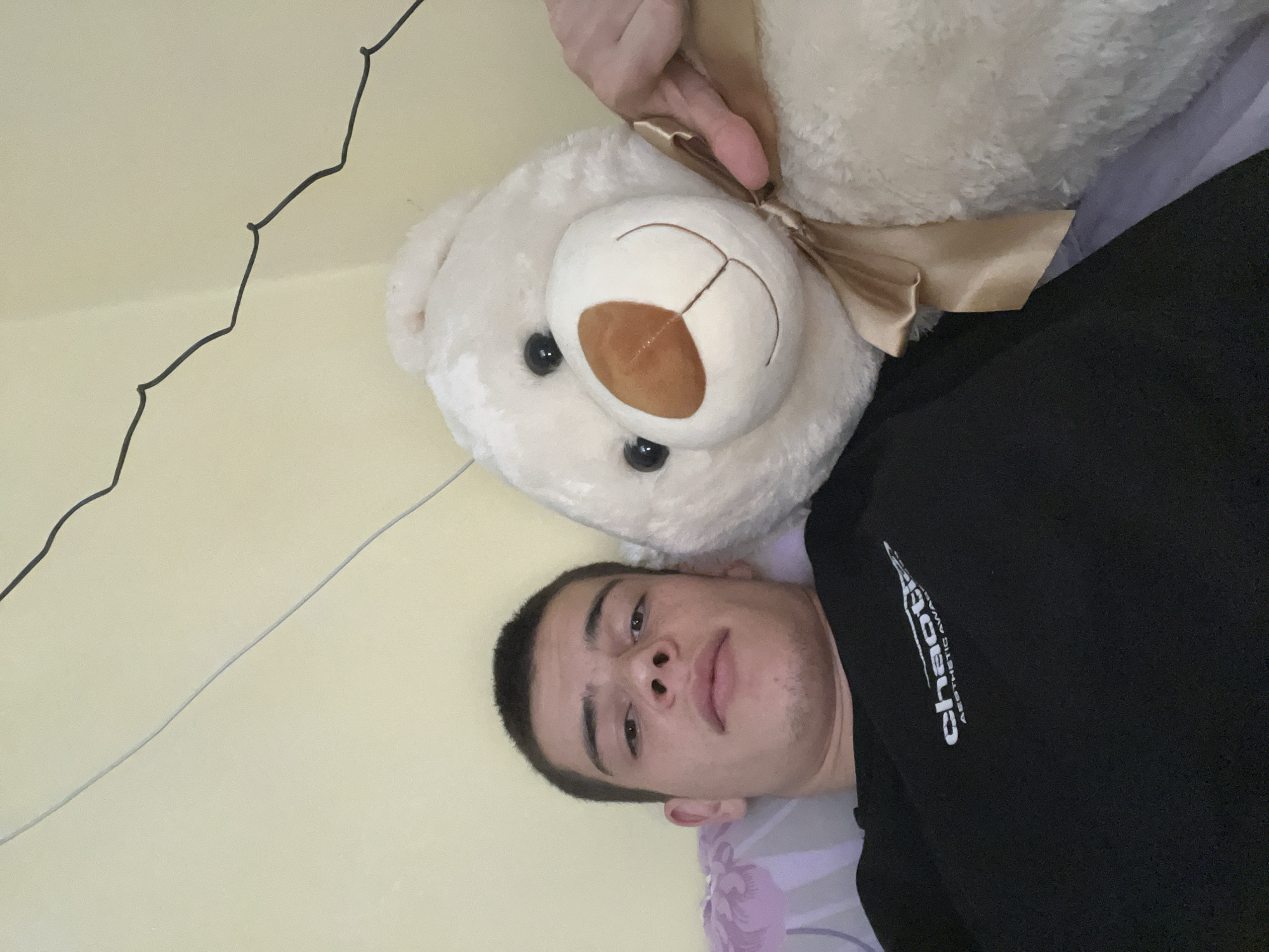 A young man lies on a bed next to a white teddy bear with a gold ribbon. The man is wearing a black hoodie with white text. The background is a light yellow wall with a black wire hanging on it. The photo was taken in a room in Plovdiv, Bulgaria.
