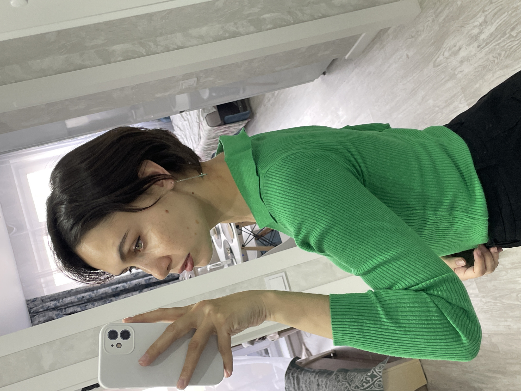 A woman with short, dark hair and fair skin is taking a selfie in front of a mirror. She is wearing a bright green, ribbed, long-sleeved shirt and black pants. She is holding a white smartphone in her left hand. The background appears to be an interior space, possibly a home, with a bed visible through the mirror and a window with curtains in the distance. The setting does not provide enough information to confirm that it is in Izhevsk, Russia.