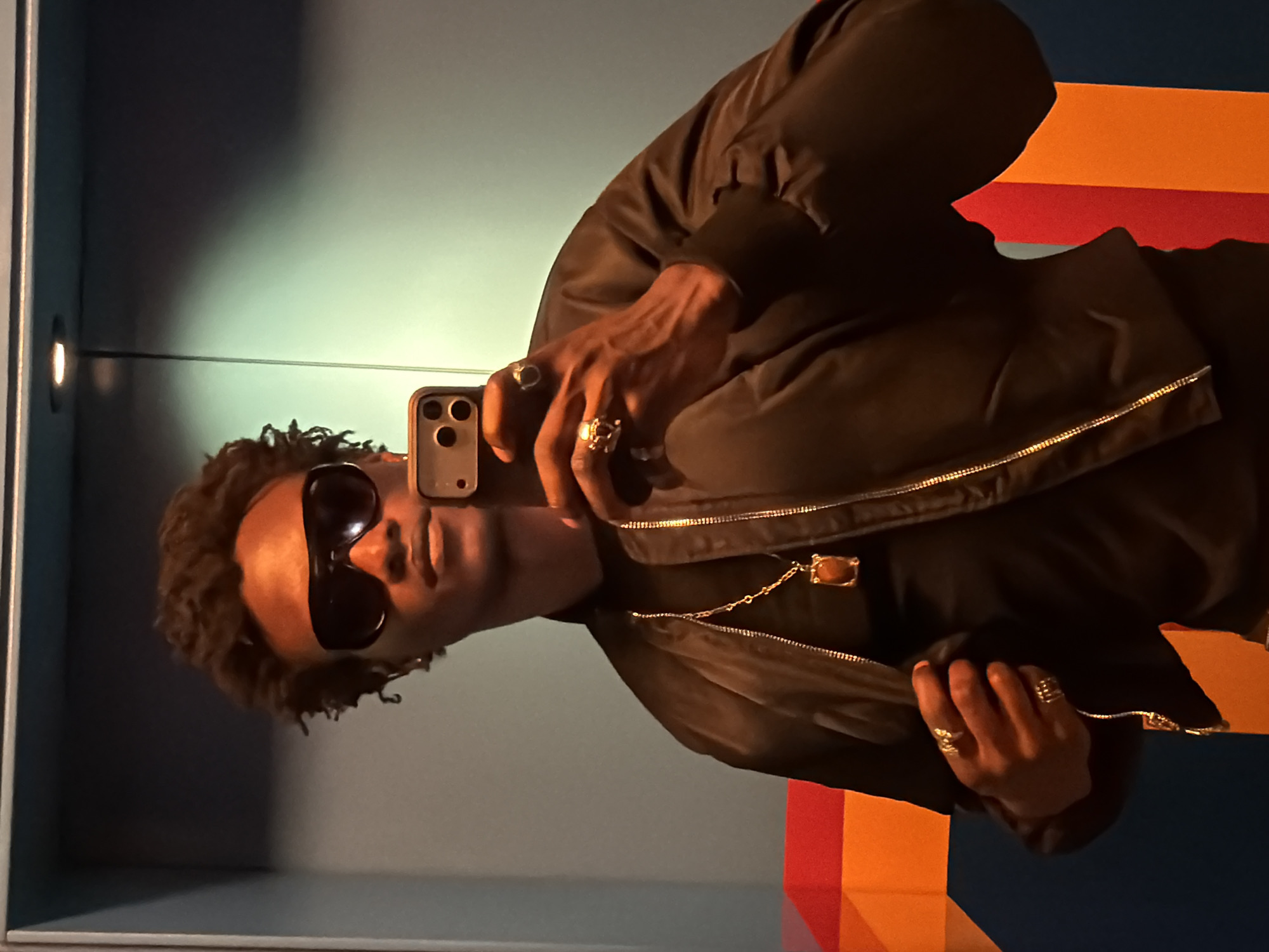 A man with dark skin is taking a selfie with his phone. He is wearing dark sunglasses, a black jacket with a gold zipper, and several rings on his fingers. He also wears a gold chain around his neck. He has dark, curly hair. The background includes a wall with a light fixture, and a section of colorful geometric shapes. The setting is indoors. It is not possible to identify a location in London, United Kingdom, from the provided image.
