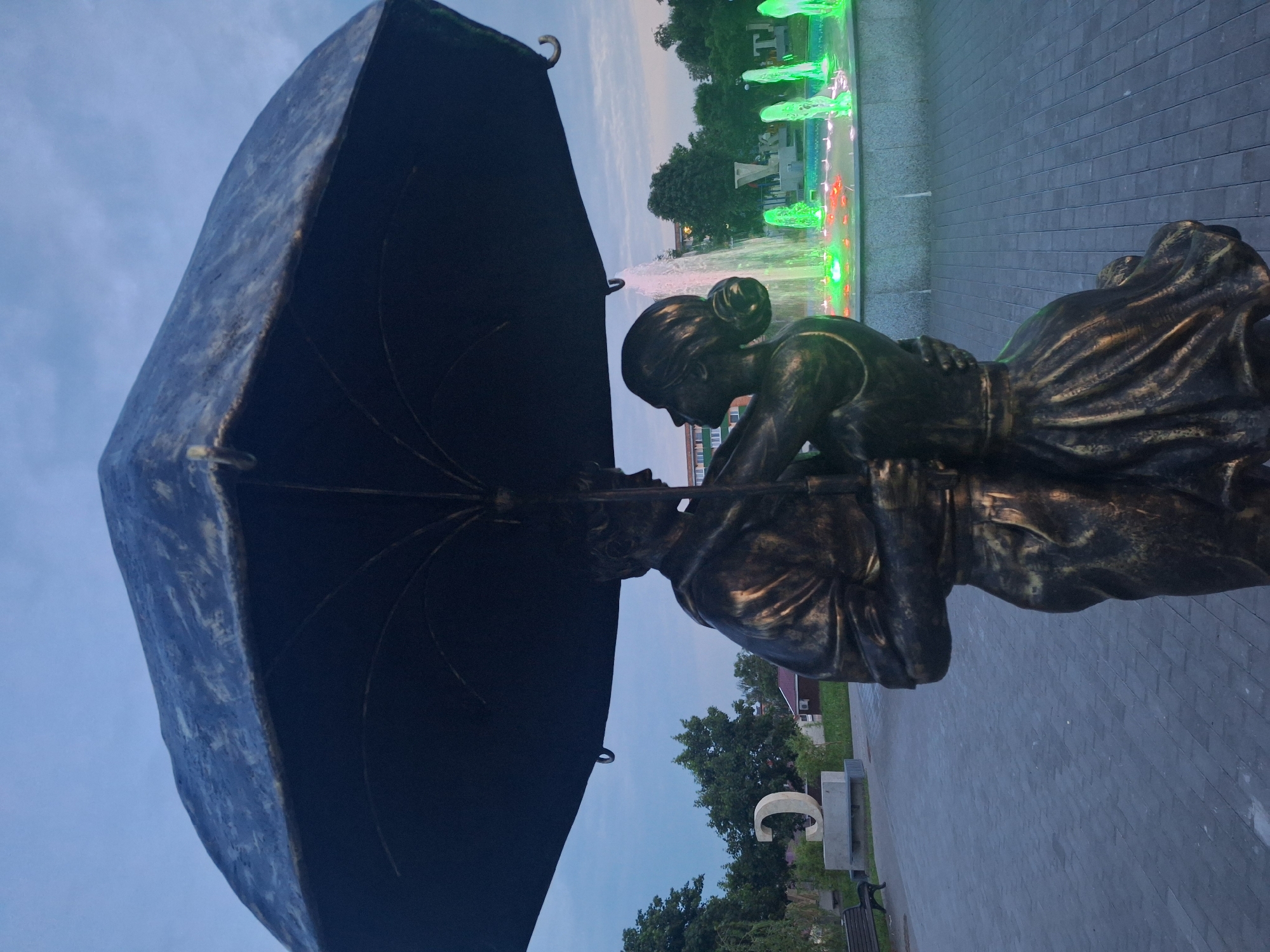 The image shows a bronze statue of a couple embracing under a large umbrella in the city of Kapyĺ, Belarus. The statue is positioned in front of a fountain with colorful lights. In the background, trees and some buildings are visible, along with a large concrete sculpture of the letter "C". The sky is overcast, indicating it may be dusk or a cloudy day. The ground is paved with interlocking stones.