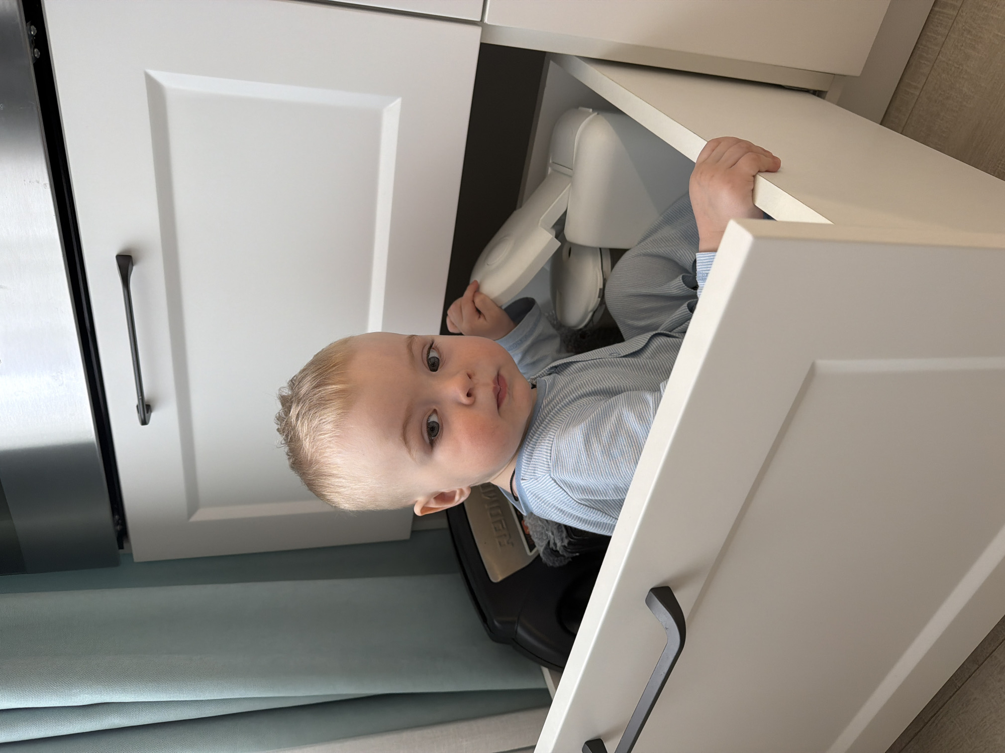 A baby is inside a partially opened white cabinet drawer. The baby is wearing a blue and white striped shirt. Inside the drawer with the baby is a white baby seat and a black object. The cabinet drawer has a black handle. A curtain is partially visible at the bottom of the image. The image was taken inside a home. The city is not discernible from the image.