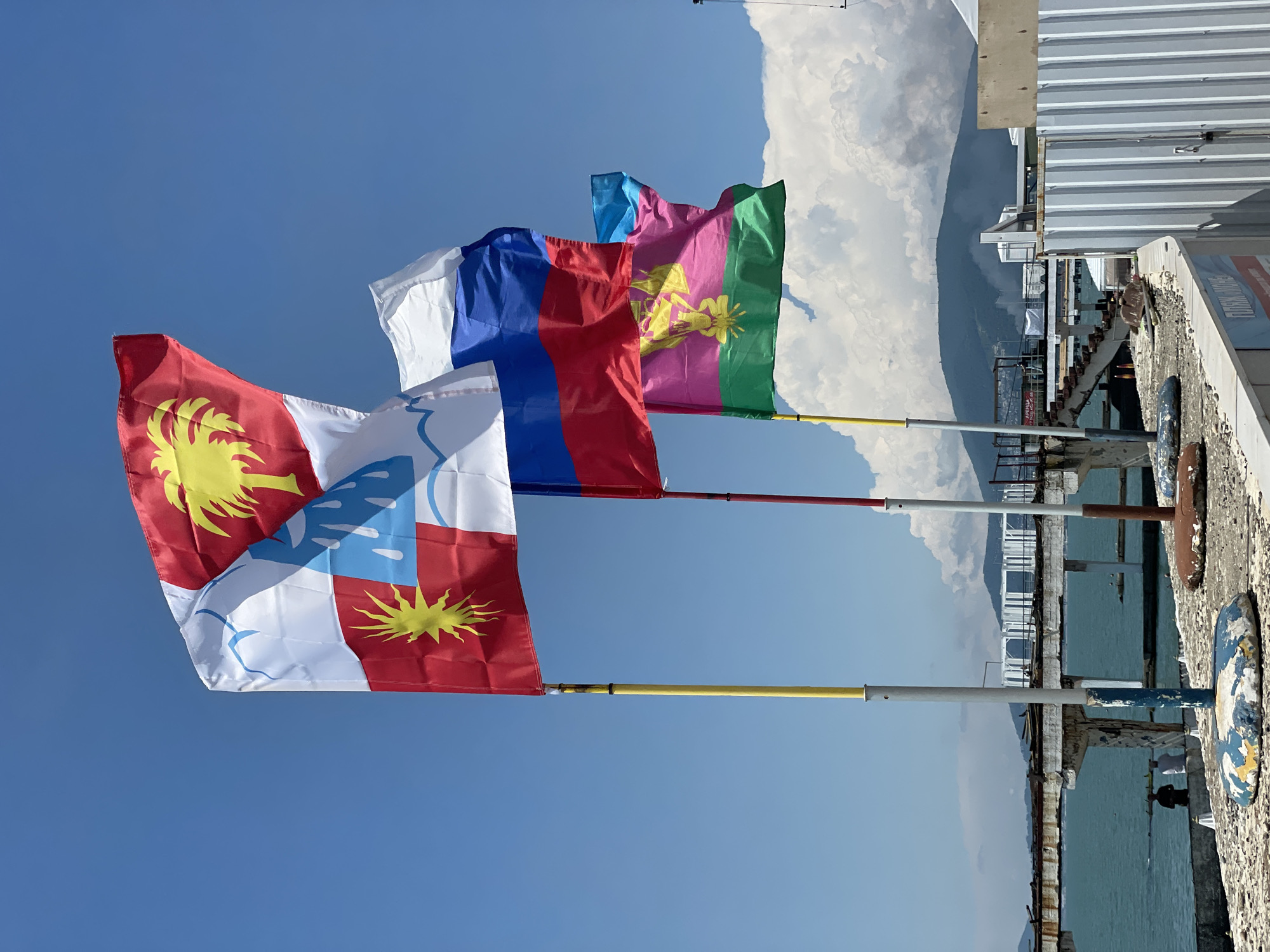 A sunny day with a clear blue sky, a few white clouds, and distant mountains serves as the backdrop for a scene featuring several flags on poles. In the foreground, three flags are prominently displayed. The topmost flag has a white field with blue wavy lines and a palm tree in a red field at the top, and a sunburst in a red field at the bottom. Below it is the flag of Russia, with its white, blue, and red horizontal stripes. The third flag, a bright pink and green one, features a yellow eagle-like emblem.
The flags are mounted on poles, some of which are yellow and red. The poles are set on a concrete platform covered in debris, with some rusted metal objects visible. To the left of the flags, a weathered pier extends into the turquoise water. Two figures, appearing to be men, are seated on a low concrete wall by the water's edge, facing away from the camera. In the distance, across the water, a white modern structure with arched roofs is visible, and beyond that, hazy mountains rise against the horizon. The overall impression is of a somewhat dilapidated seaside area under a bright sky.
This scene is likely located in Sochi, Russia, given the presence of the Russian flag and the distinctive coastal landscape with mountains characteristic of the region. The weather and the presence of beach-related infrastructure, albeit in a state of disrepair, further suggest a Russian Black Sea resort town like Sochi.