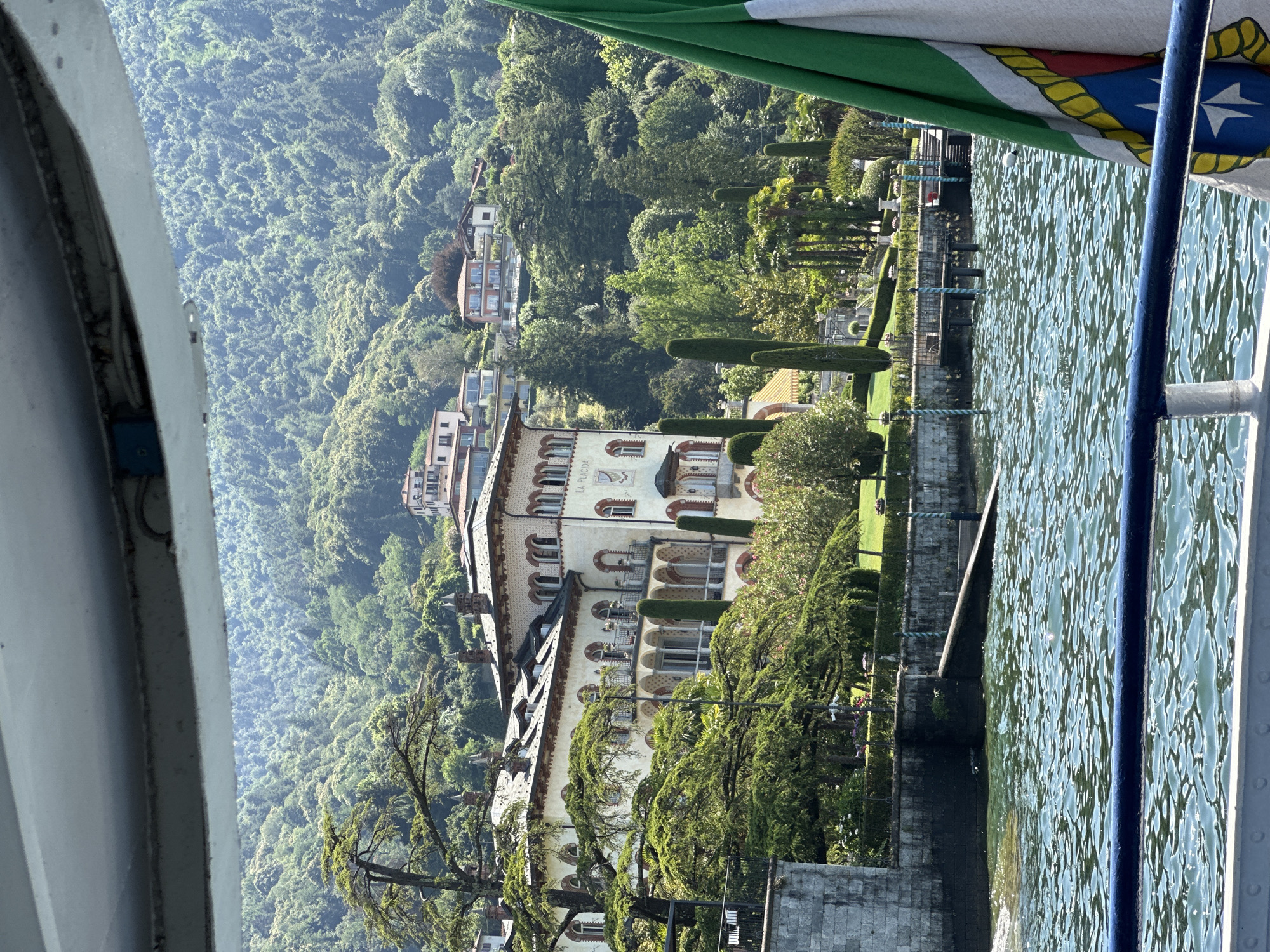 The image captures a view from a boat on a body of water, looking towards a large, ornate building situated on a hillside. The building, with its cream-colored facade and red-tiled roof, features numerous arched windows and balconies. Lush green trees and manicured gardens surround the building. In the background, the hillside is densely covered with trees, and several houses are visible nestled among them. The water in the foreground is choppy, reflecting the bright sunlight. A portion of an Italian flag, with its green, white, and red colors, is visible in the lower right corner. The scene suggests a scenic location, possibly a lakeside villa or estate. The inscription "LA PLACIDA" is visible on the side of the building. The location is identified as San Giovanni, Italy.