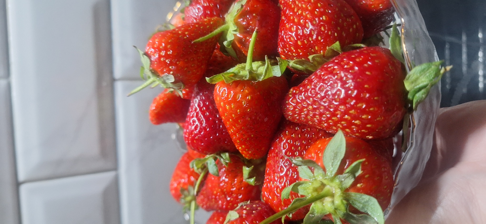 A close-up shot reveals a clear plastic container filled with ripe, red strawberries with green stems and leaves. The container appears to be held by a hand on the right side of the frame. The background includes a white tiled wall on the left and a darker surface or object on the right, partially obscured. There is no indication of location in Moscow, Russia.