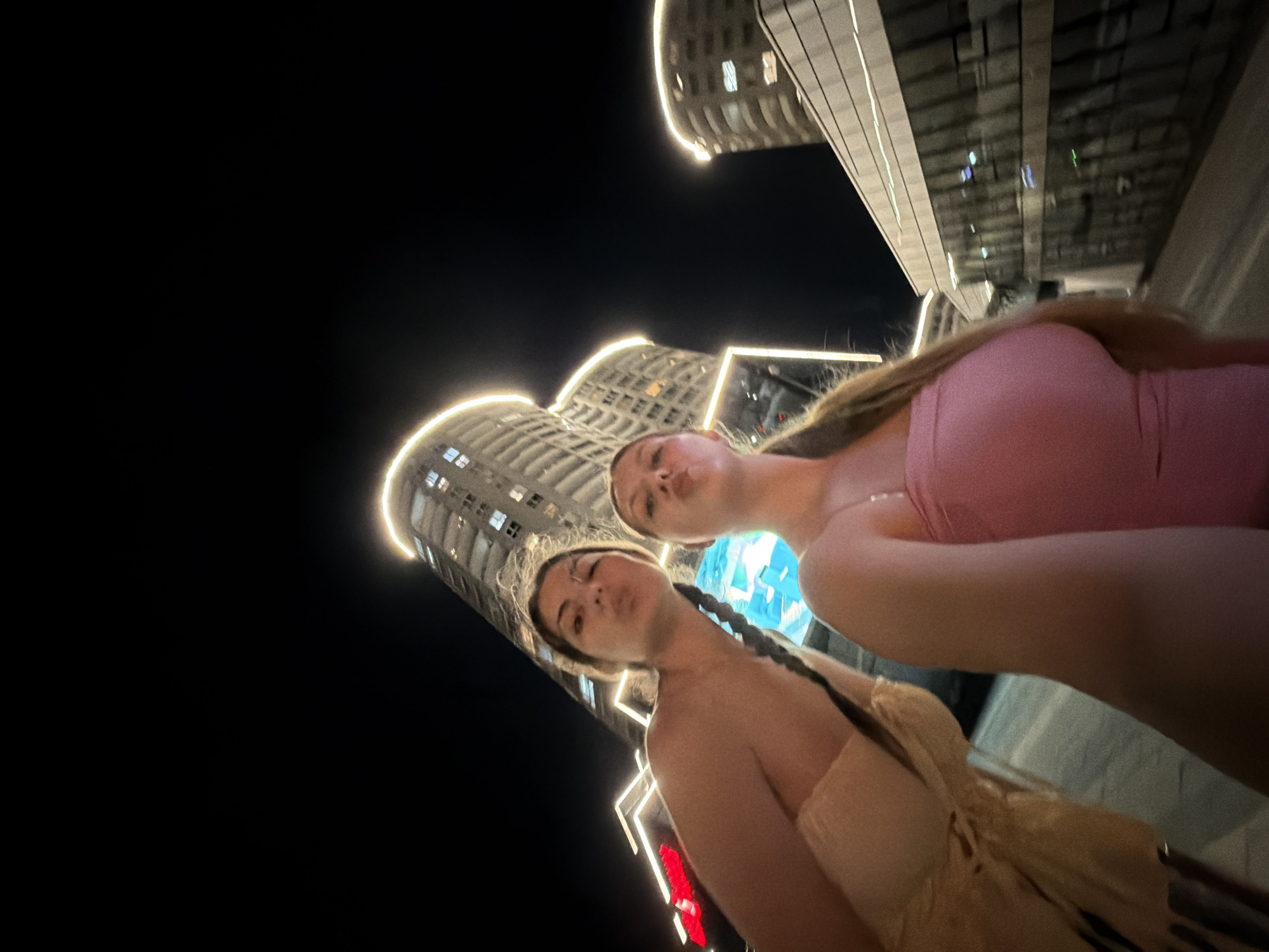 The image captures two young women posing for a low-angle photo at night in Burgas, Bulgaria. Both subjects are looking towards the camera with playfully puckered lips. The woman in the foreground on the left has dark hair, partially styled in braids, and wears a light brown or beige strapless top. The second woman, positioned slightly behind and to the right, wears a pink top and has long light brown hair.
Behind the women, the scene is dominated by striking modern architecture. Several tall, multi-storied buildings with curved facades are visible against the dark night sky. These structures are prominently outlined with bright, warm-toned horizontal strip lighting, highlighting their unique design. The artificial illumination from the buildings and potentially other urban lights provides the primary light source for the scene. A blurred red glow, possibly from a neon sign or other urban lighting, is visible in the lower-left background. No legible text is visible. The atmosphere is that of a lively urban night, suggesting a clear evening for an outdoor photograph.