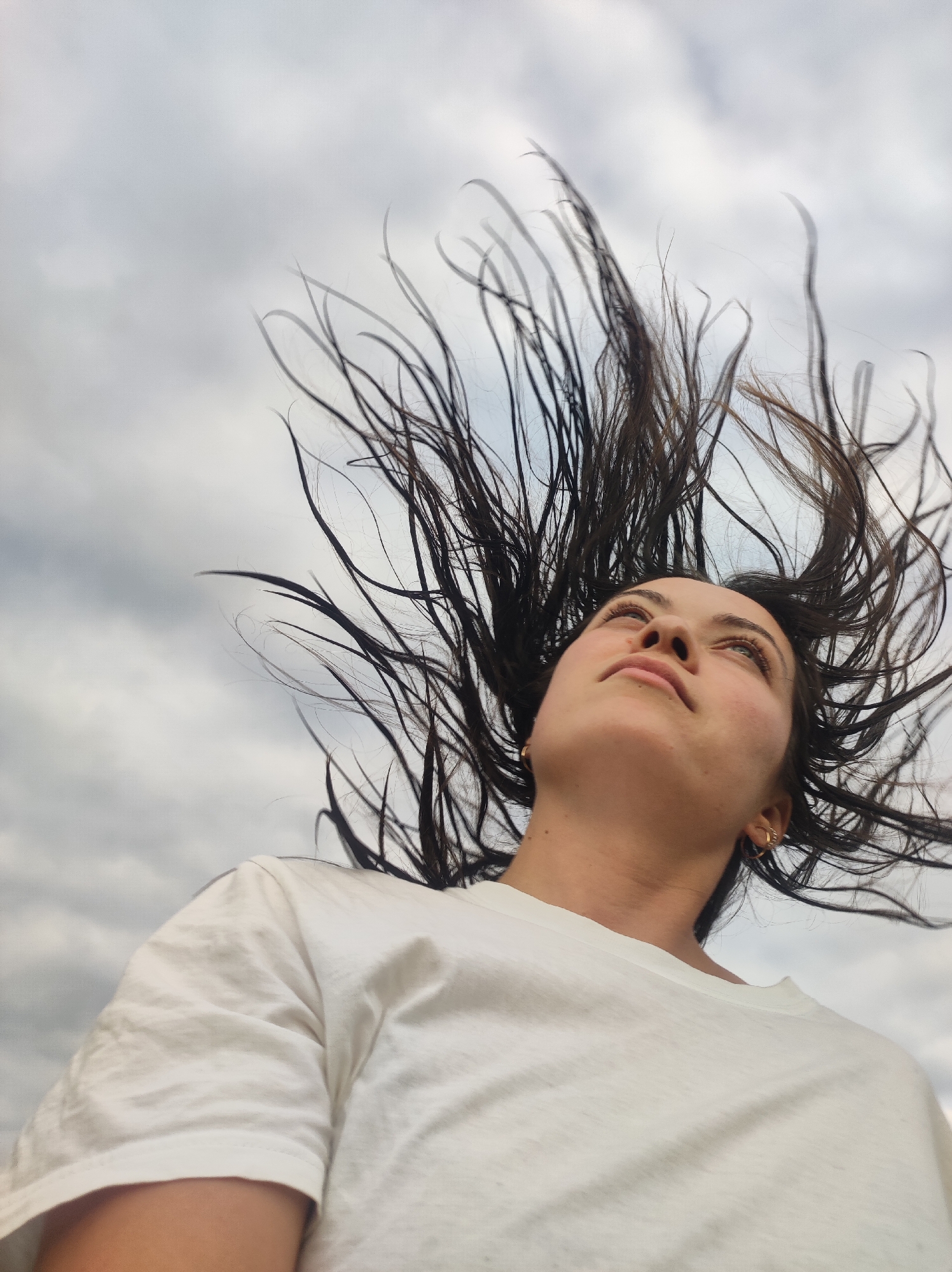 A young adult female with dark hair and light eyes is centered in the frame, looking upward with her head tilted back against an overcast sky. Her dark hair is dramatically fanned out around her head, extending upwards and outwards, suggesting motion from a head flip or strong upward wind. She wears a plain white short-sleeved t-shirt and small gold hoop earrings on both ears. The image is captured from a low-angle perspective. The background consists entirely of a cloudy, light grey sky. The scene is located in the city of Фоки, Russia.