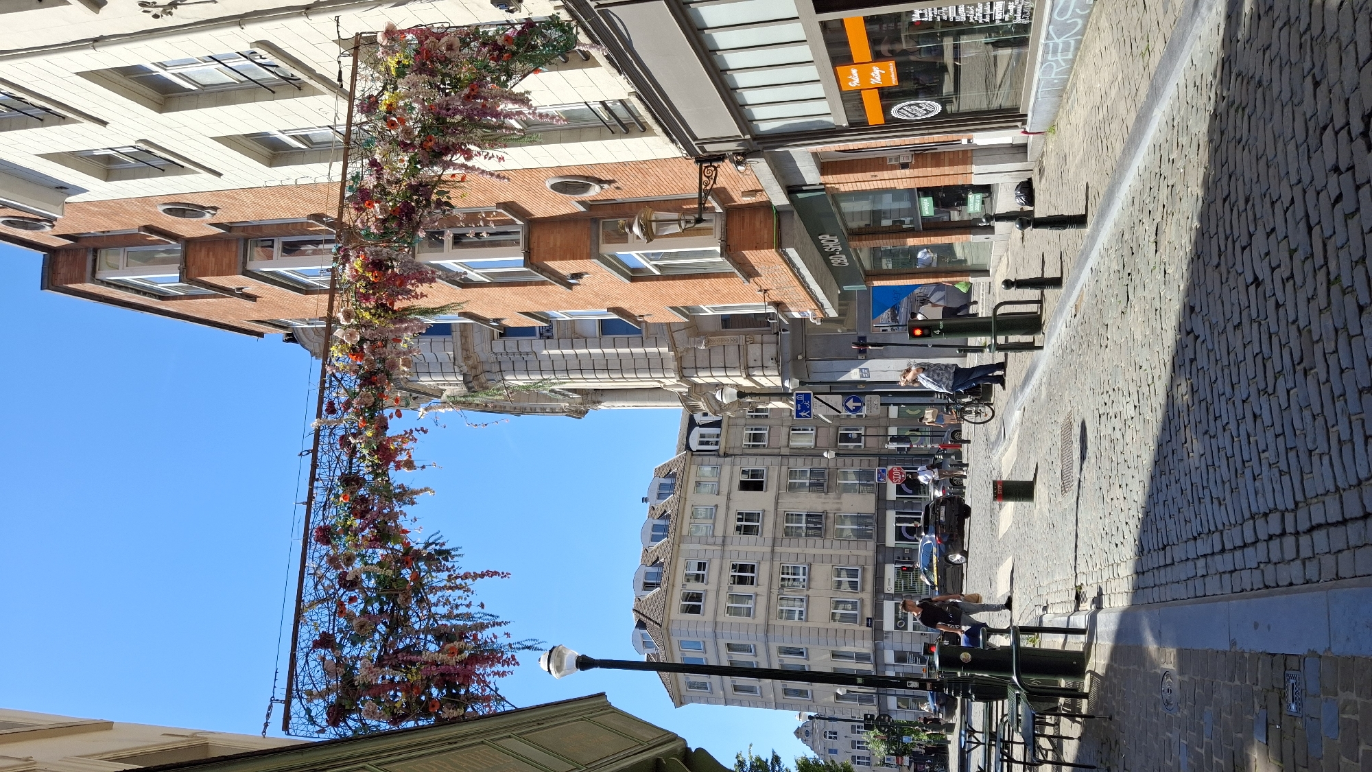 The image depicts a sunlit urban street scene on a clear day, likely in a European city given the architecture and cobblestone paving. A decorative floral archway made of various colorful flowers hangs across the street, spanning between buildings.
On the left, a building with a green awning and a modern facade partially frames the view. A lamppost stands on the sidewalk. Further down the street, a multi-story building with numerous windows and a mansard roof is visible. A black car with its headlights on is parked on the street, and a few pedestrians are walking.
In the center and right of the image, a brick building with multiple windows and balconies is prominent. Below it, a store with a green awning has the text "CBD SHOP" on its facade. Next to it, another store features large windows with signage, including the word "Vintage."
The street is paved with cobblestones, and shadows from the buildings and the floral archway are cast across it. Several black bollards are positioned along the sidewalk. Traffic lights are visible, one of which is red.
A woman in a blue and white patterned top and jeans is standing near a bicycle and looking up and to her right. A man in a dark t-shirt and light-colored pants is walking towards the left, carrying a blue bag. Another man in a white shirt is walking away from the camera.
The sky is a clear, vibrant blue. The overall impression is of a lively, well-maintained city street.