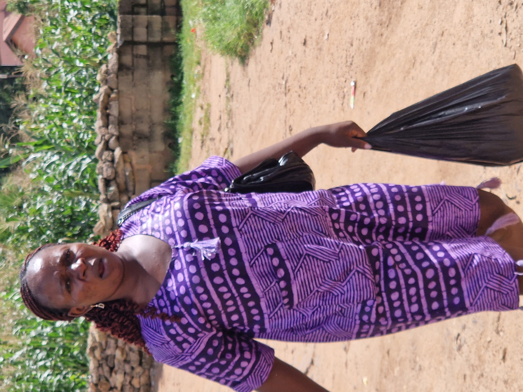 A woman with dark skin and braided hair is walking towards the viewer's left, holding a black plastic bag in her right hand. She is wearing a purple and black patterned garment that appears to be a romper or a two-piece outfit. The garment has circular and geometric patterns. She is also wearing a black purse over her left shoulder. The background shows a dirt path, some green vegetation, a stone wall, and a building in the distance. The scene appears to be outdoors in a rural or semi-rural area, possibly in the city of Jos, Nigeria.