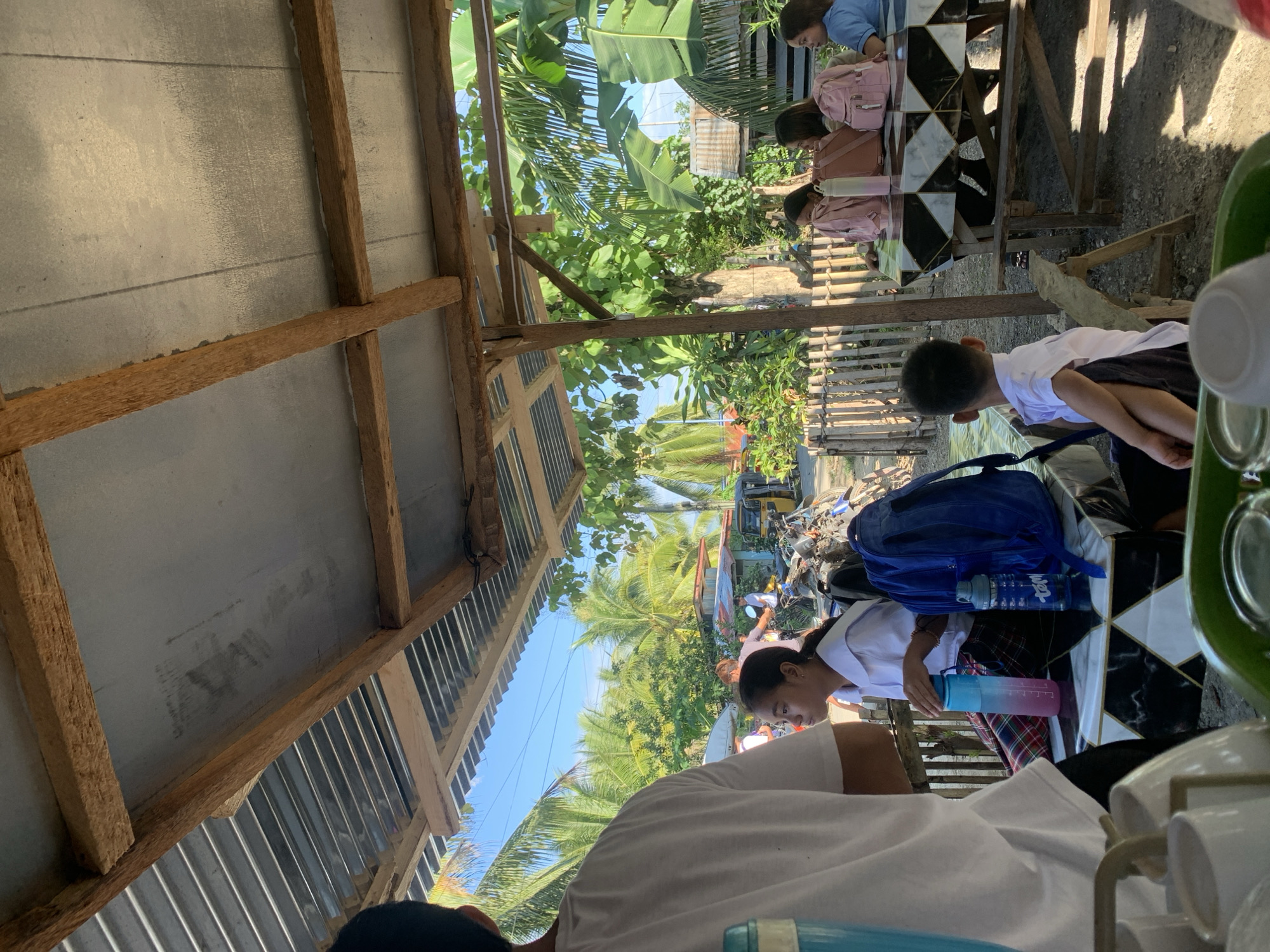 In Sulop, Philippines, under a corrugated tin roof supported by wooden beams, a scene unfolds outdoors. A young girl in a white school uniform and a plaid skirt sits at a table, holding a pink and blue water bottle. Next to her are two other girls, one wearing a pink backpack, and another with a similar pink backpack. A young boy in a white shirt and dark shorts sits at another table, facing away from the camera. In the background, palm trees and lush green foliage indicate a tropical environment. A motorcycle and a trishaw are visible, suggesting transportation typical of the area. The overall impression is of a casual gathering in a semi-rural, possibly roadside, setting.