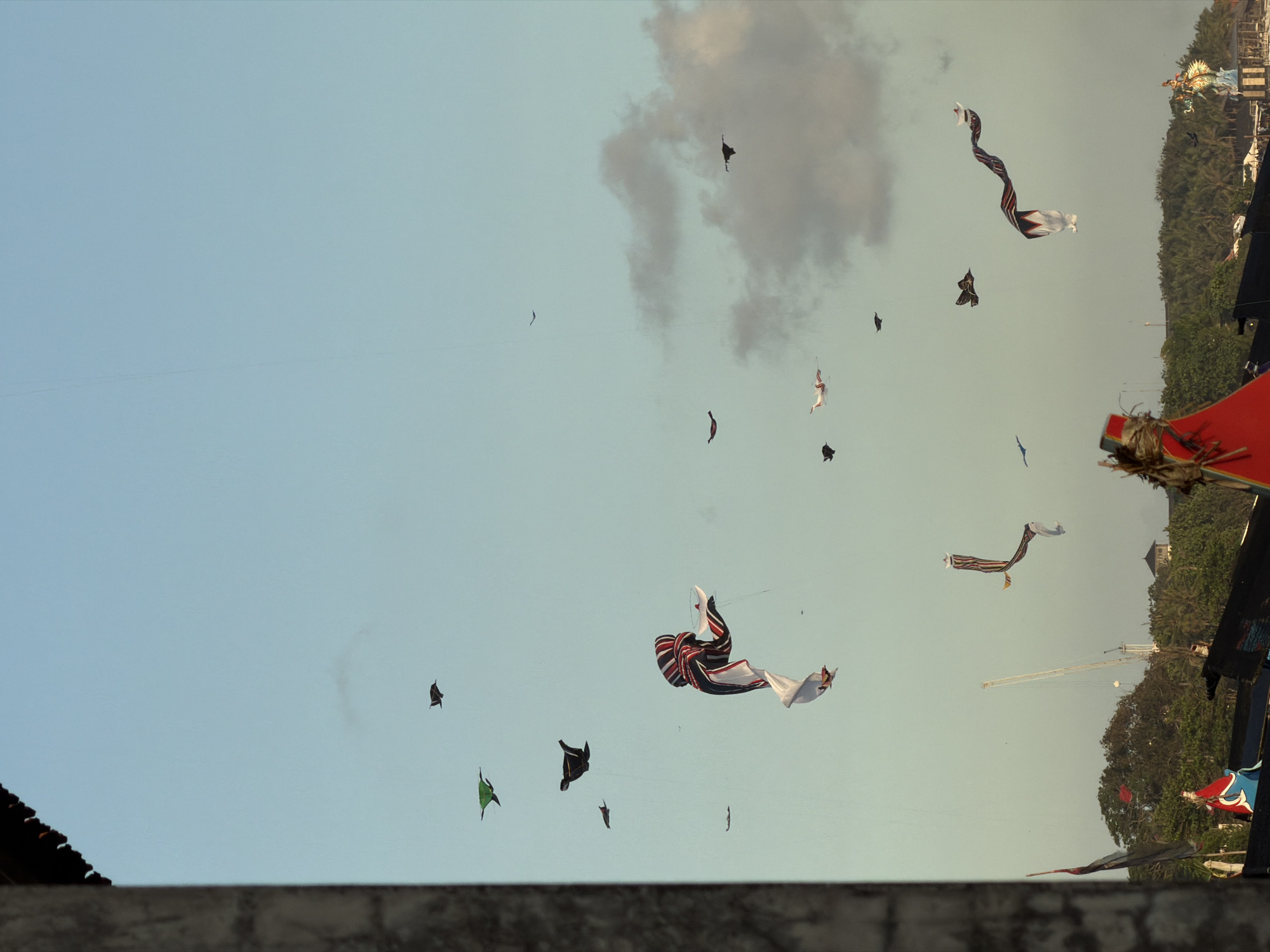 The image depicts a sky filled with various kites. The location is Mengwi, Indonesia, indicated by the presence of Balinese kites. Numerous kites of different shapes, sizes, and colors fill the sky, including traditional Balinese kites. Some kites are black, green, or white, while others have intricate patterns. A large cloud is present in the upper right portion of the image. The sky is a light blue color. In the background, a tree line and some buildings can be seen. The photo appears to be taken from a slightly elevated position. Parts of a building are visible at the bottom of the frame.
