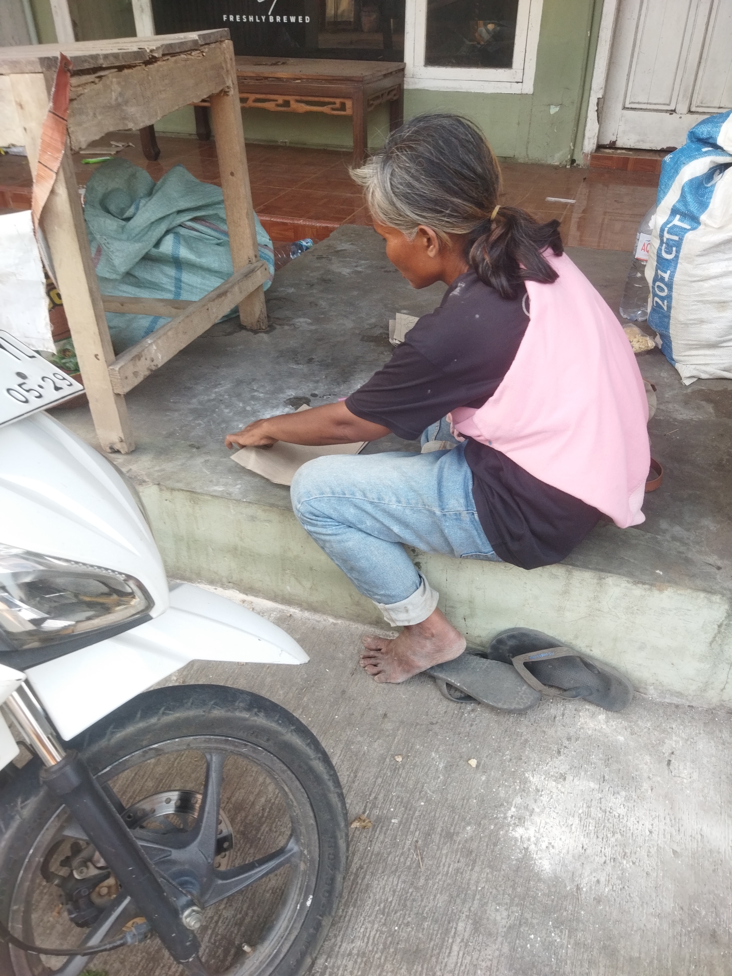 An adult person with grey and black hair tied in a ponytail, wearing a black t-shirt and a pink vest, and blue jeans, sits on concrete steps with their legs crossed. The person's feet are bare, and one foot rests on a pair of dark-colored flip-flops. In front of the person, on the steps, is a piece of brown paper being worked on by their hands. To the left of the person is a white motorcycle. To the right of the person are two large white bags with blue markings. The environment appears to be an outdoor area with a concrete floor and a wall in the background. The image was taken in the city of Banjar, Indonesia.