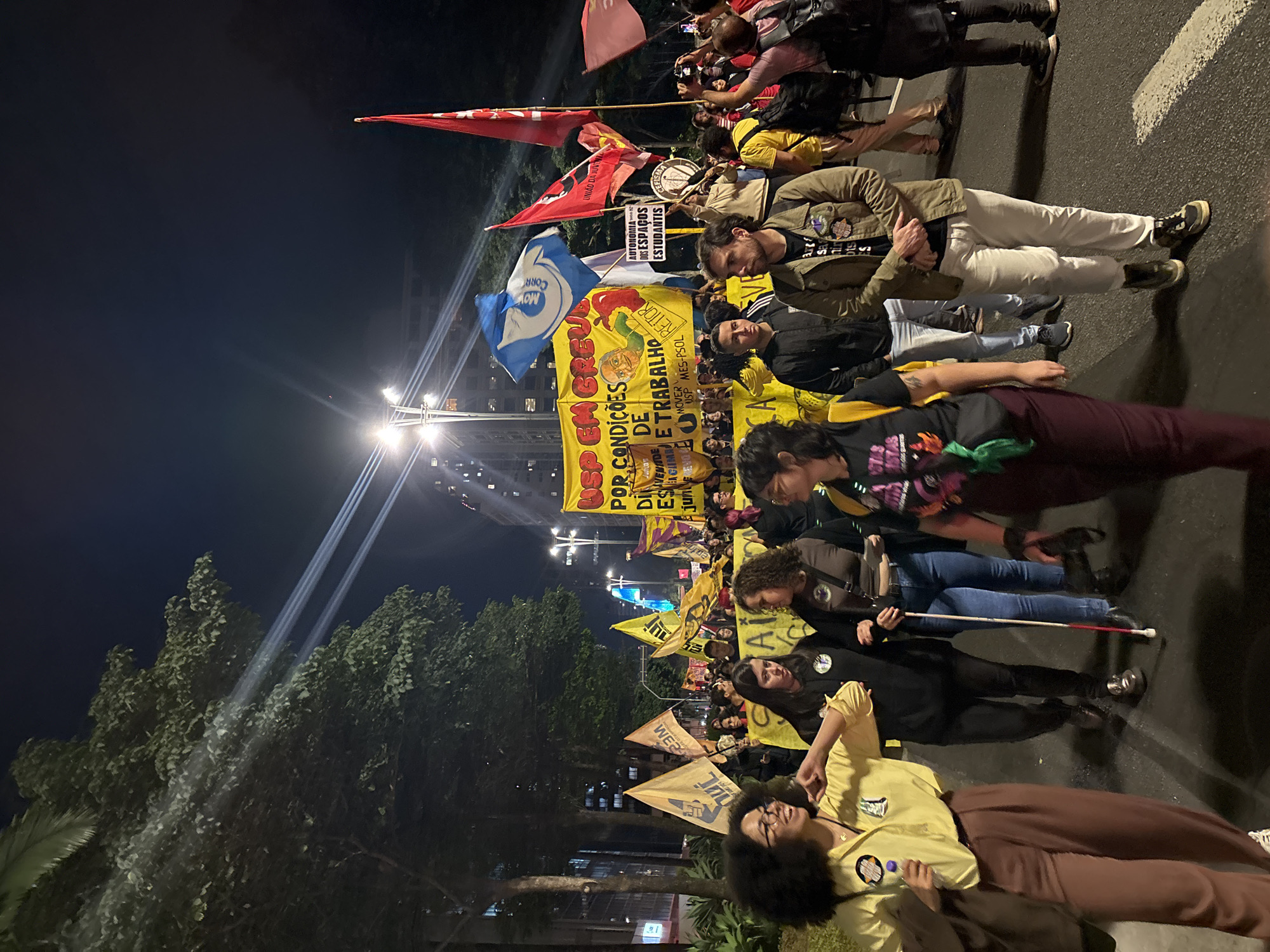 A nighttime street scene shows a political protest in São Paulo, Brazil. A group of people are visible carrying signs and banners. Flags and banners with political messages are displayed, along with flags, indicating it is an organized demonstration. The banners and signs include text such as "POR CONDIÇÕES DE TRABALHO" and "AUTONOMIA DOS ESPAÇOS ESTUDANTIS." The street is illuminated by streetlights. Trees and buildings are visible in the background, placing the event in an urban setting. The protesters are interacting with each other and marching forward.