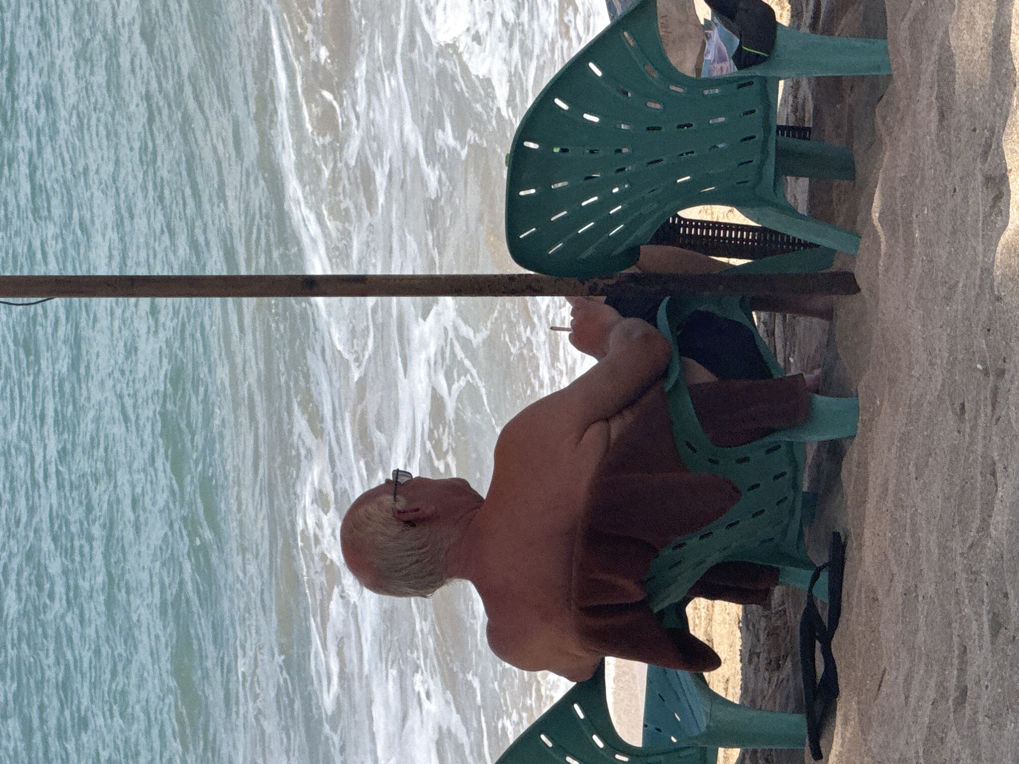 An elderly Caucasian male, bald with white hair around the ears, sits in a green plastic beach chair facing the ocean in Kuta, Indonesia. He is shirtless and wearing dark sunglasses. A brown towel is draped over the back of his chair. He holds a lit cigarette in his right hand. Next to him, to his left, is another green plastic beach chair, empty. To his right, a large metal pole, possibly part of a beach umbrella, stands vertically. The ocean is visible behind him, with waves gently rolling in. The ground is sandy beach.