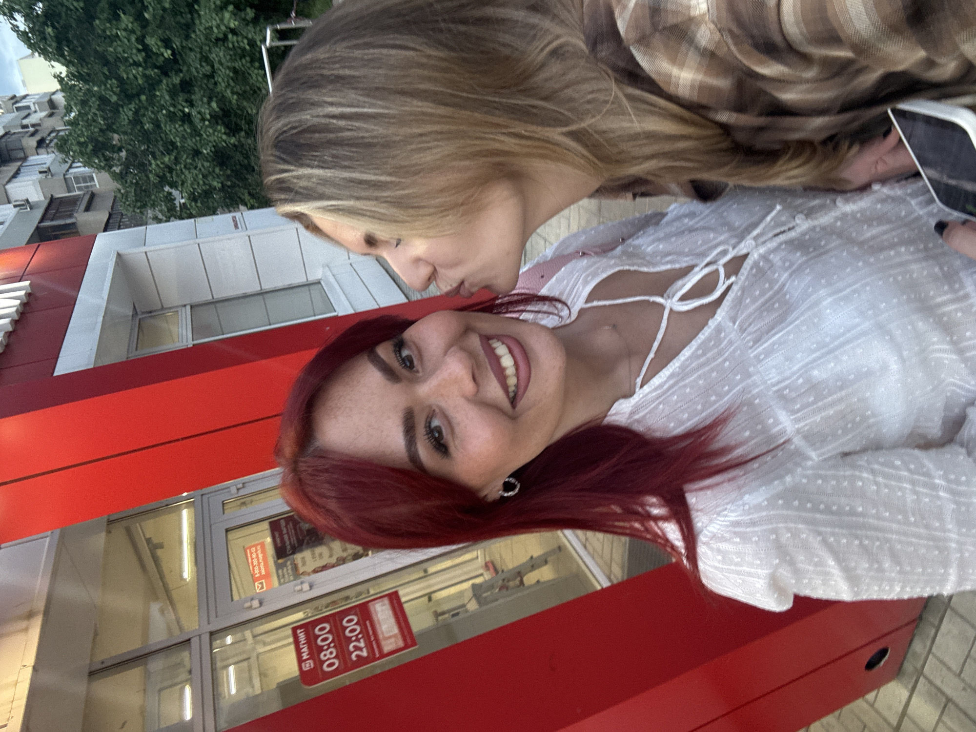 Two women are captured in a selfie outside a store. The woman in the foreground on the left has bright red hair, freckles, and is wearing a white textured blouse and hoop earrings. She is smiling at the camera. The woman on the right has blonde hair and is leaning in to kiss the other woman's cheek. She is wearing a plaid shirt. Behind them, a building with red and white accents is visible, featuring a storefront with a glass door. A sign on the door displays store hours. The scene appears to be in an urban environment, with trees and apartment buildings visible in the background.