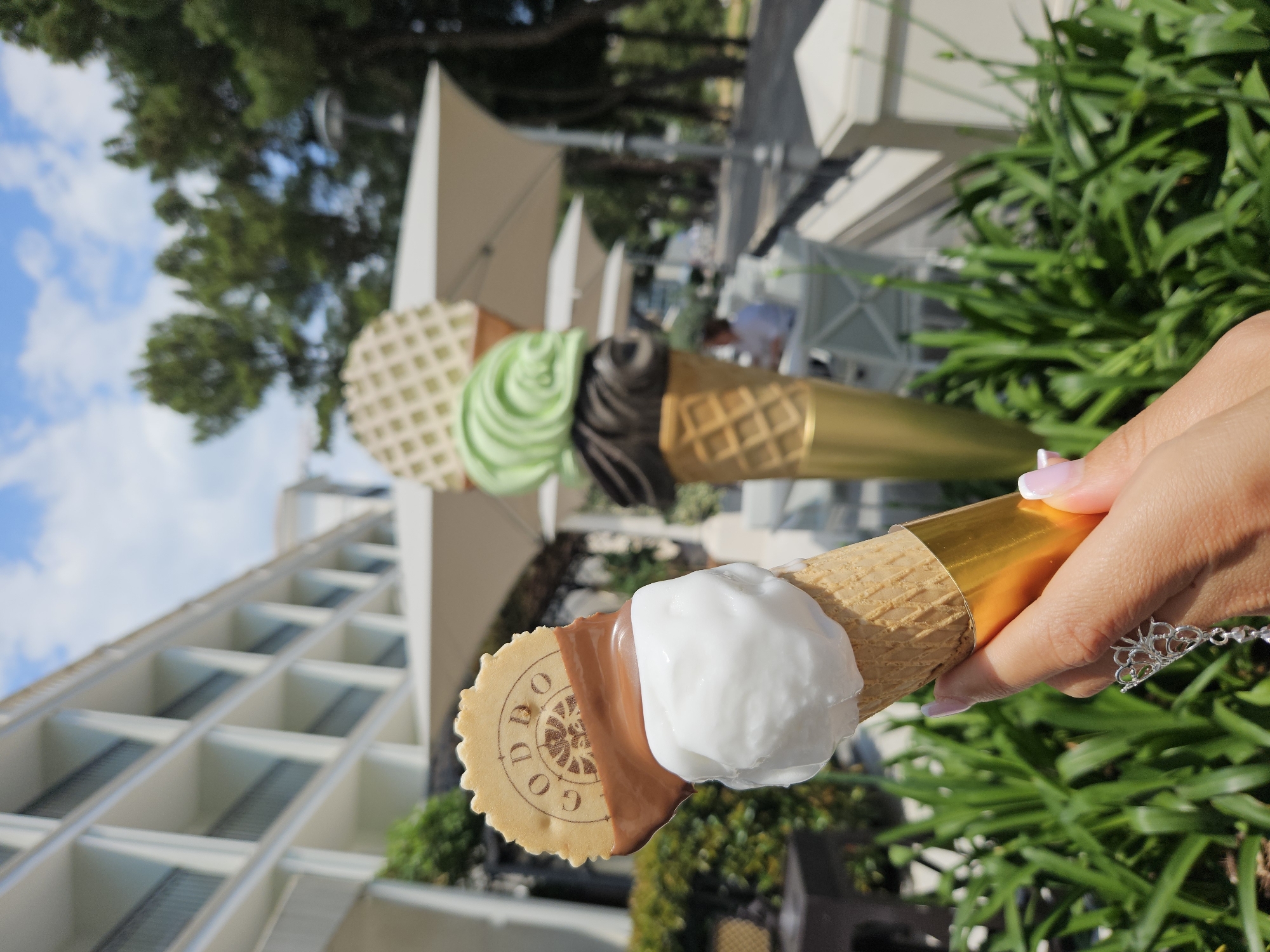A close-up, outdoor shot shows a person holding an ice cream cone with white ice cream and chocolate topping, with a decorative wafer reading "GODDBOO" on it. The cone is wrapped with gold foil. In the background, there are two large ice cream cone sculptures, one with green and brown "ice cream" and the other possibly gold, standing in front of white tables with umbrellas. In the distance is a white, multi-story building with a grid-like facade. Green foliage is visible around the ice cream cone and in the background.