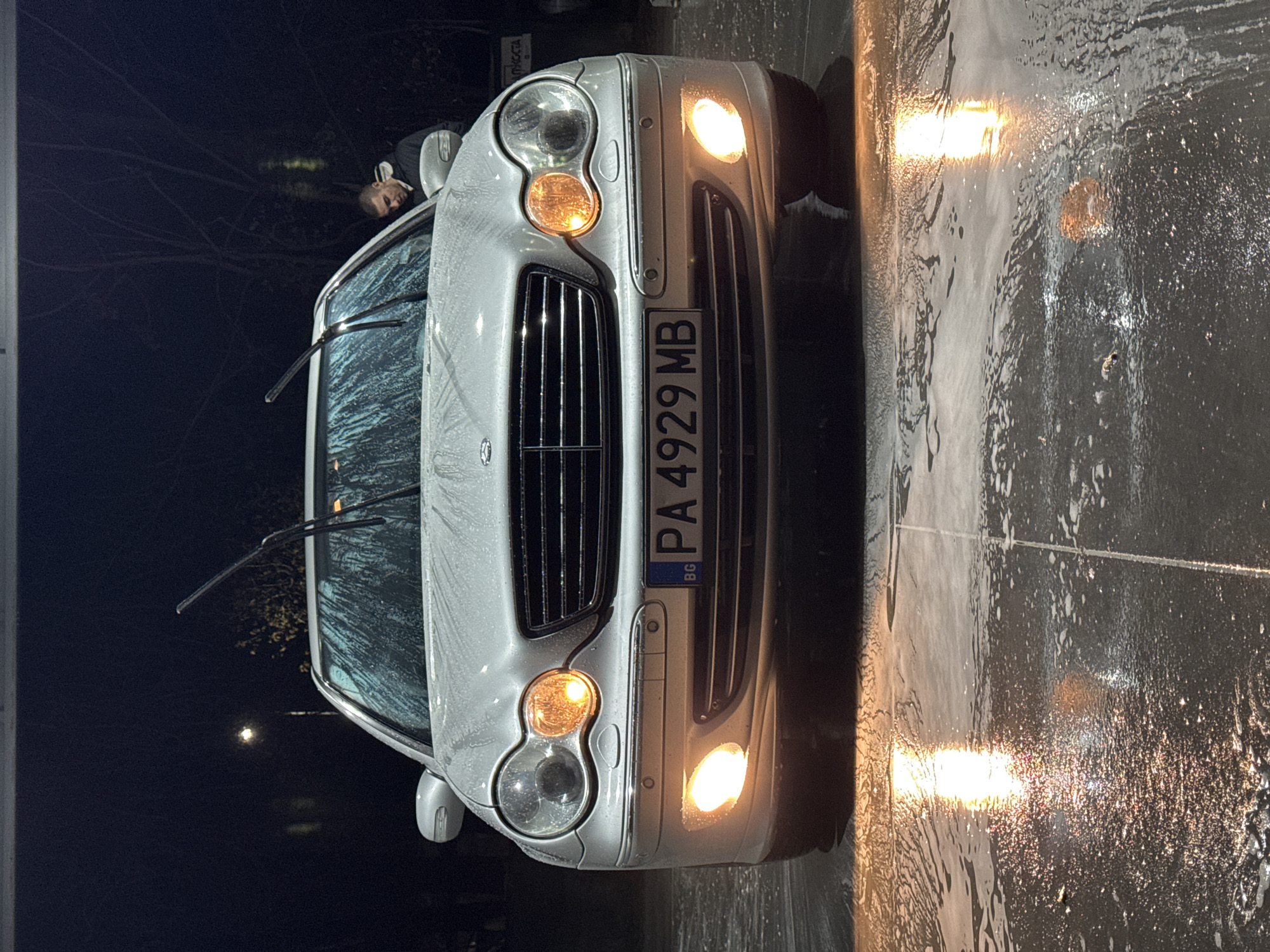 The image shows a silver Mercedes-Benz car at a car wash location. The car's license plate reads "PA 4929 MB" and includes "BG" indicating Bulgaria. The car is covered in soap suds, particularly on the windshield. Its headlights and turn signals are illuminated. A person is partially visible near the upper left corner of the car, near the driver's side. The background is dark, suggesting it is nighttime. The wet ground surface reflects the light from the headlights. The city is Plovdiv, Bulgaria, based on the "BG" license plate identifier.
