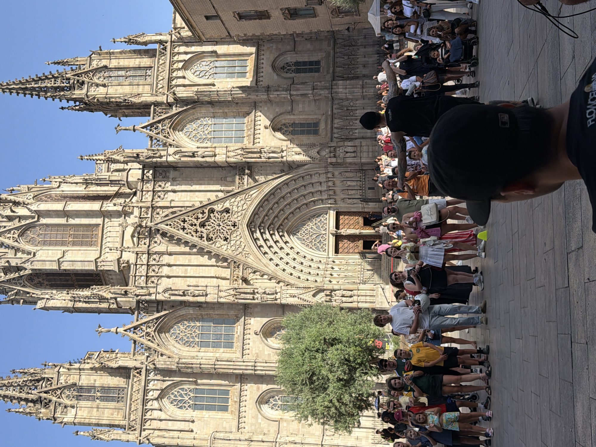 The image captures a lively street scene in front of the Barcelona Cathedral in the Gothic Quarter of Barcelona, Spain. A large, ornate Gothic cathedral with intricate stonework, arched windows, and towering spires dominates the background. The sky is clear and blue.
In the foreground, a crowd of people of various ages is gathered. Many are looking towards the cathedral or at a street performer who is facing away from the camera with arms outstretched, wearing a black tank top and a black beanie. The performer has tattoos on their arms and back. In front of the performer, a person wearing a black baseball cap is facing the performer, partially obscuring the view of some of the crowd.
The crowd consists of tourists and locals, dressed in casual summer clothing. Some are holding up their phones, likely taking pictures or videos. Children are present, and one is being pushed in a stroller. Among the people are a young boy in a yellow t-shirt and shorts, standing near the front, and a young girl with blonde hair in a colorful outfit to his left. To the right of the boy, a young man in a white t-shirt stands holding a white helmet.
A large green tree stands to the left of the cathedral entrance. The ground is paved with cobblestones. Several yellow cones are placed in the square, possibly marking off an area or for a performance. The atmosphere appears to be festive and engaging, with a street performance drawing the attention of the onlookers.