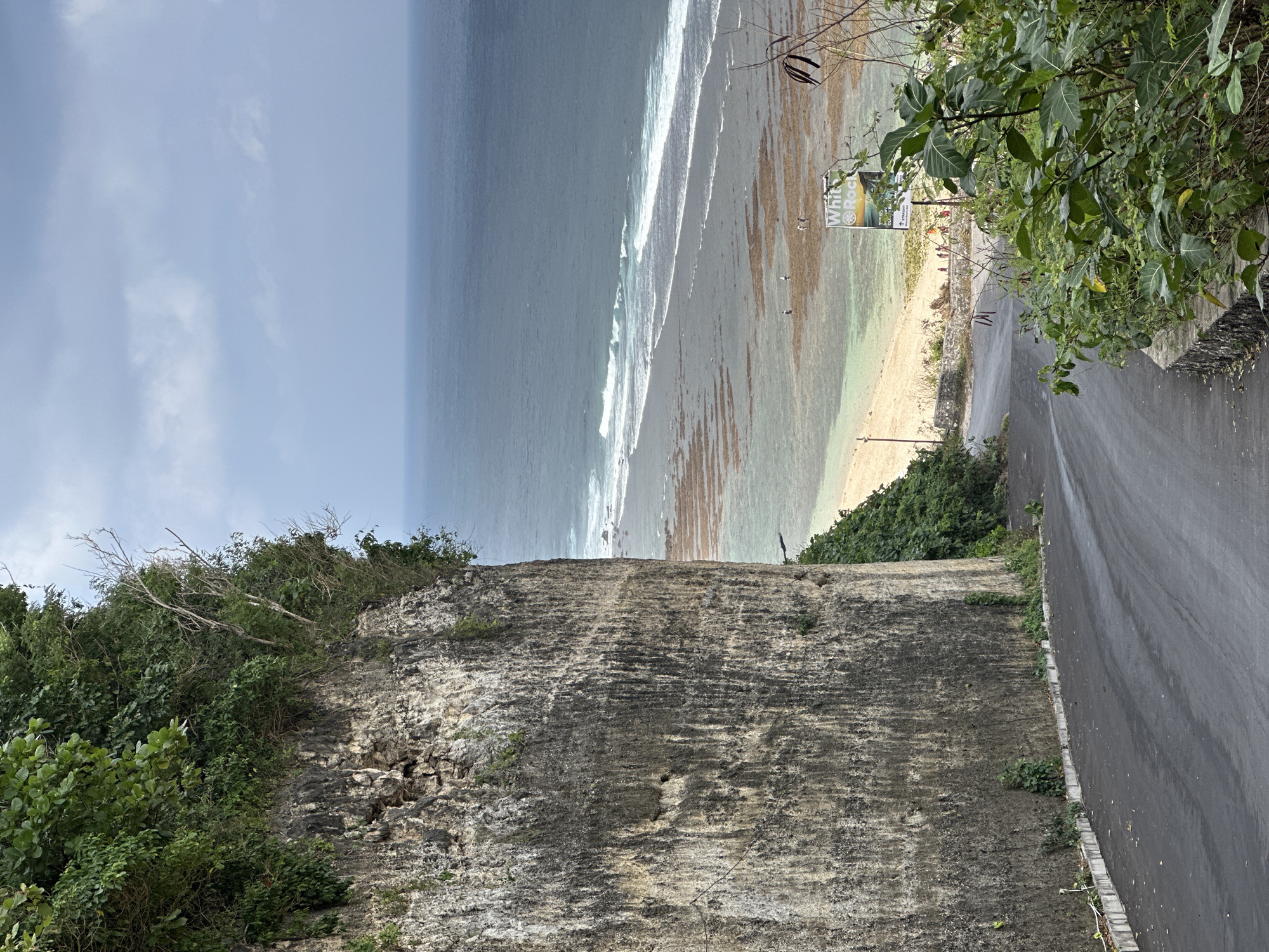 The image depicts a winding asphalt road descending towards a beach with calm turquoise waters and gentle waves breaking on the shore. The road is bordered by a steep, rocky cliff face on the left, covered in lush green vegetation at the top and smaller plants at its base. On the right, more foliage lines the road. In the distance, the ocean stretches to the horizon under a cloudy sky. A sign with the text "White Rock" and graphics is visible near the beach. Several small figures, likely people, are scattered on the sandy beach and in the shallow water. The scene appears to be a scenic viewpoint overlooking the coastline in Kuta Selatan, Indonesia.