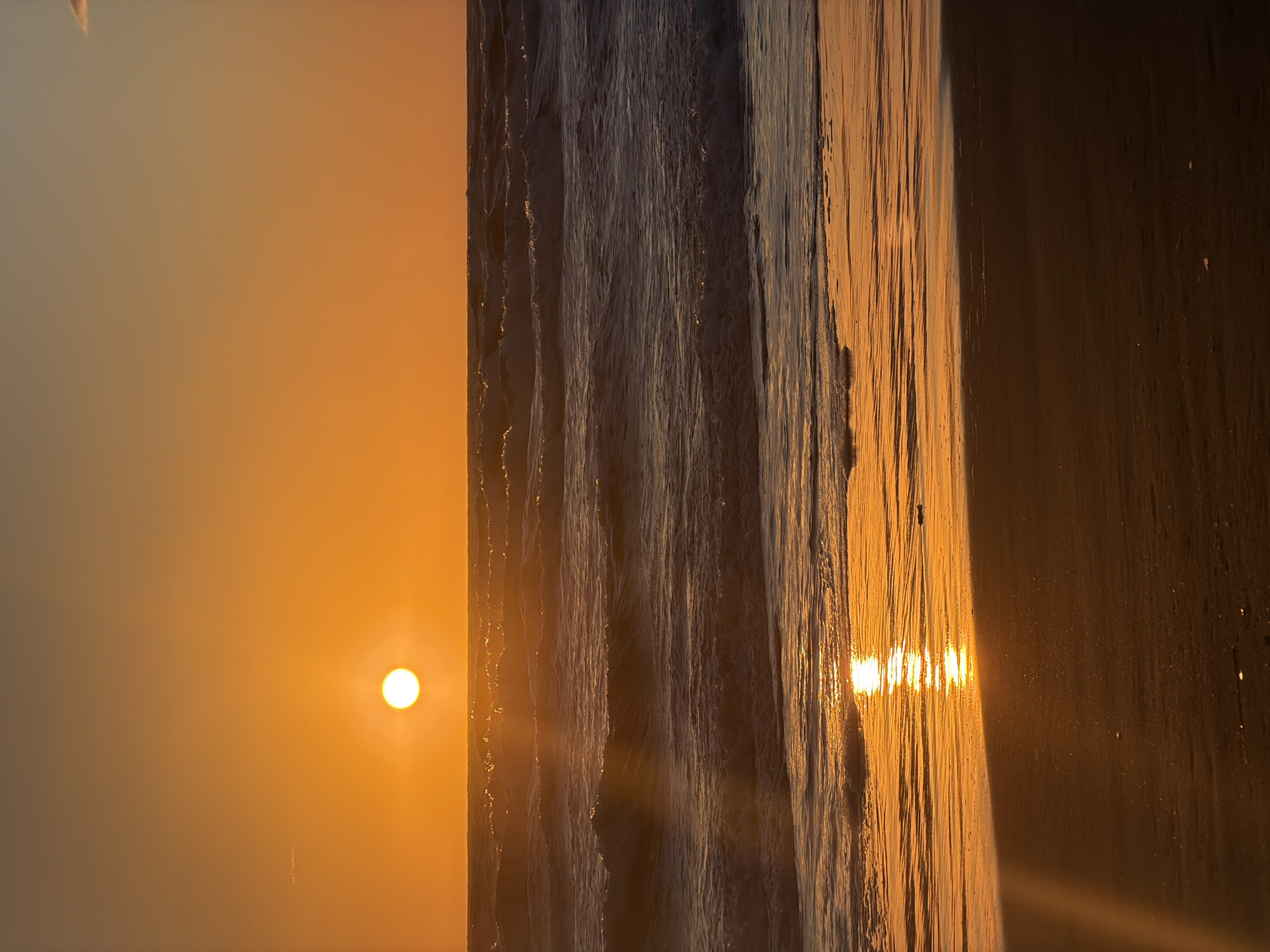 The image shows a sun over the ocean in Castagneto Carducci, Italy. The sun is low in the sky on the left side of the image. Waves are on the right side of the image, they are reflecting the sunlight.