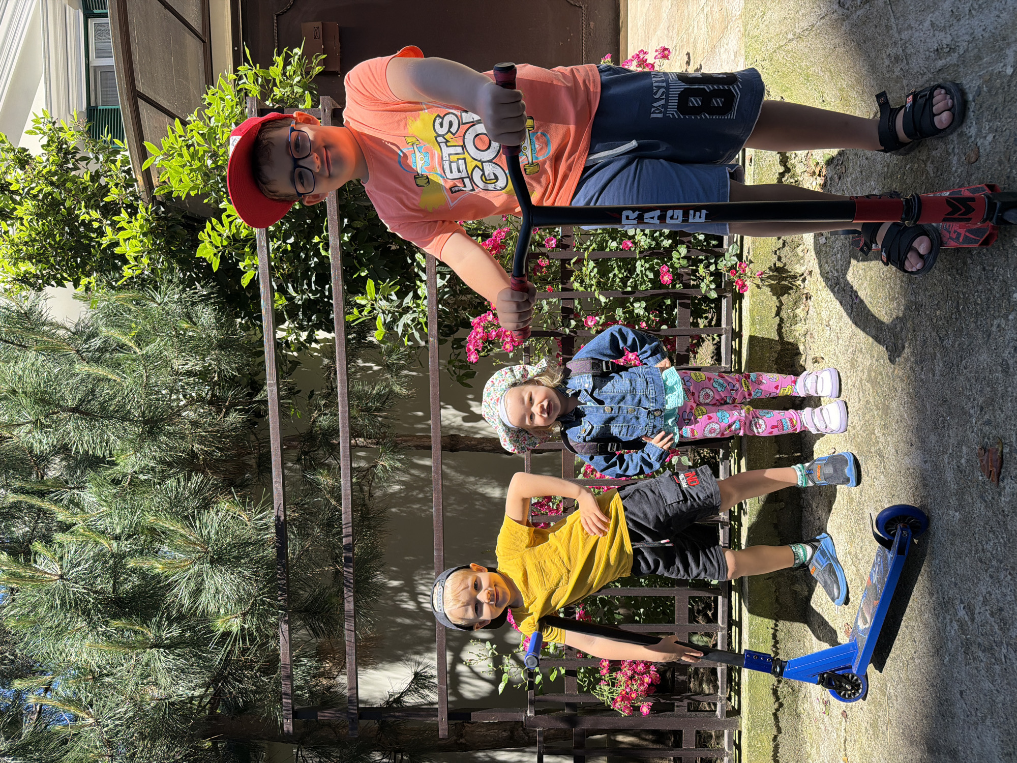 An outdoor, eye-level, full-shot photograph captures three young children posing with scooters on a sunny day in what appears to be a paved yard or garden in Sochi, Russia. The image is taken in portrait orientation but displayed sideways.
On the right, a boy with fair hair, wearing a red cap, glasses, an orange t-shirt with "LET'S GO!" printed on it, and blue shorts with "FASTER" visible on one leg, stands holding a black and red scooter with "RAGE" written on its stem. He is looking at the camera with a smile.
In the center-left, a young girl with light hair covered by a floral sun hat, wearing a denim jacket, and brightly patterned leggings, stands smiling towards the camera.
On the bottom, another boy with fair hair wearing a dark cap, a yellow t-shirt, and dark shorts with "PRESS ONE" visible, poses with a blue scooter. He also smiles at the camera, with one hand on his hip and the other on the scooter's handlebar.
The background features green foliage, including pine needles and flowering bushes with small pink blossoms, behind a dark metal fence. Part of a building with white walls and a window is visible in the upper background. Strong shadows indicate bright, direct sunlight, suggesting a midday or early afternoon setting. All three children appear happy and cheerful.