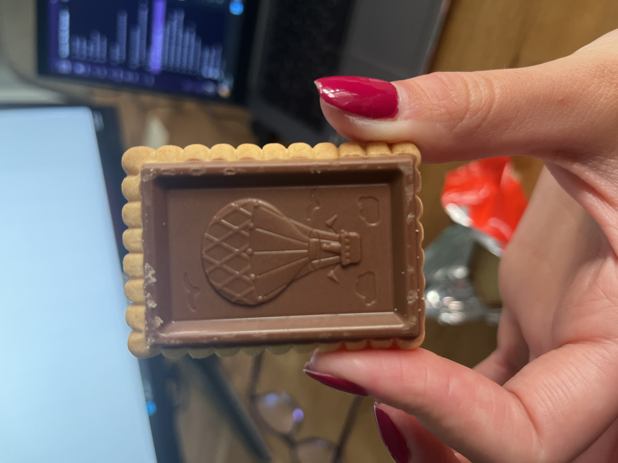 A person's hand, with bright red nail polish on the thumb and index finger, holds up a rectangular biscuit. The biscuit features a scalloped edge and is topped with a layer of milk chocolate. Embossed on the chocolate is a clear design of a hot air balloon, alongside two small heart shapes to its right.
The background is blurred, focusing attention on the biscuit. To the top left, a screen displays an abstract pattern of vertical bars in purple and blue, possibly a data visualization or audio waveform. Below this, another light-colored screen is partially visible, suggesting a laptop or tablet. Further right in the background, a crumpled red and silver object, resembling packaging, can be seen, along with a wooden surface. The scene takes place indoors, likely at a desk or table. No discernible text is visible on the biscuit or in the blurred background elements.