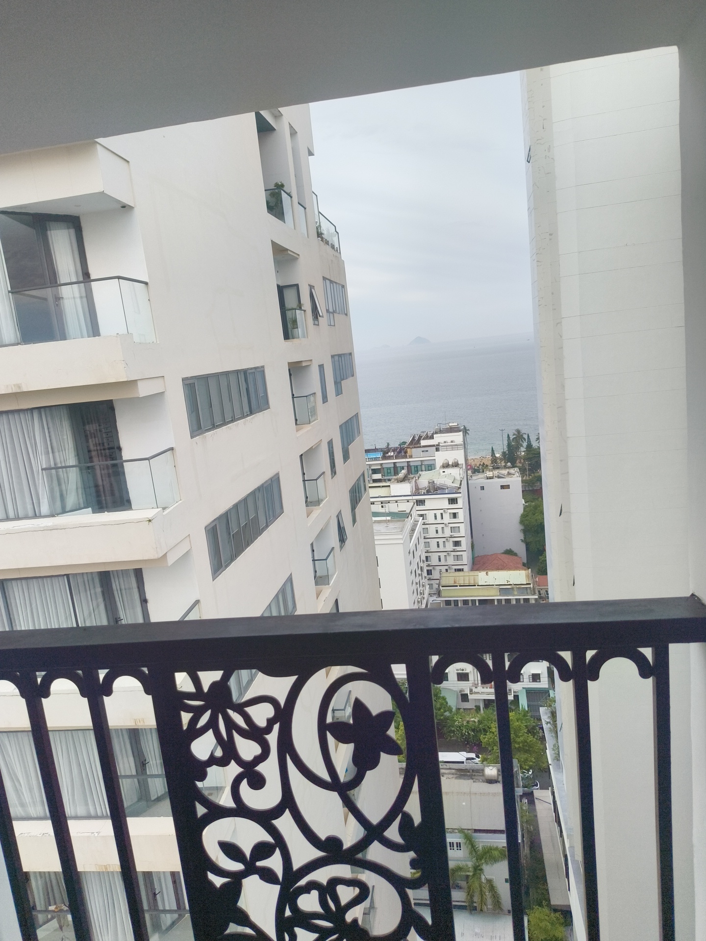 This media file captures a view from a balcony overlooking a coastal city, likely Nha Trang, Vietnam. The scene is dominated by modern white buildings of varying heights, suggesting a developed urban environment. In the foreground, a black metal railing with decorative scrollwork frames the view. Beyond the buildings, a body of water, presumably the ocean, stretches towards a hazy horizon where distant islands or landmasses are faintly visible.
The architecture of the buildings features multiple balconies, large windows, and clean, geometric lines. Some balconies are furnished with glass railings, while others appear to have simpler designs. The windows are mostly covered with sheer white curtains, giving the impression of residential or hotel accommodations. The overall color palette is muted, with predominantly white buildings contrasting with the grey sky and the blues and greys of the sea.
The weather appears overcast, with a diffused light suggesting it might be morning or late afternoon, or simply a cloudy day. There are no clear indications of specific activities or events taking place, presenting a static view of the cityscape. The presence of palm trees and other lush greenery interspersed between the buildings hints at a tropical or subtropical climate.
No people are clearly visible in the image, and the focus is on the architectural and natural landscape. The overall mood is one of quiet observation, offering a glimpse into the urban and coastal scenery of a resort-like destination. There is no visible text in the image.