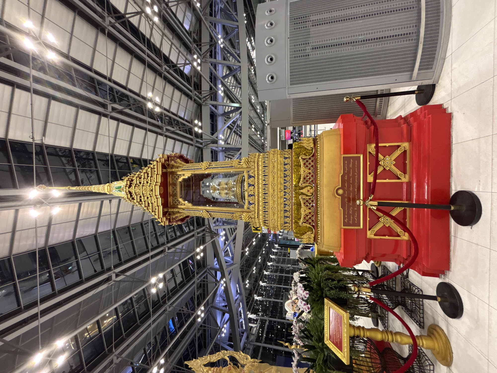 The image shows a large, ornate golden structure with a red base, featuring intricate carvings and embellishments, located indoors. The structure is likely a shrine or a monument, possibly a Buddha statue encased within. It is roped off with red velvet ropes and stanchions. To the left, there's a golden stand with a framed document. To the right of the shrine, a large grey metal unit with circular vents and a perforated front panel is visible, possibly an air conditioning unit or filtration system. The background reveals a modern airport terminal with a high, metallic ceiling structure with numerous lights, and a row of windows with a cityscape or outdoor view beyond. The flooring is light-colored tile. The lighting suggests it is daytime, but the indoor environment is brightly illuminated by artificial lights. The scene is devoid of people. This structure is located in the Suvarnabhumi Airport, which is located in Bang Phli, Samut Prakan province, Thailand, near the city of Nong Prue Subdistrict, Thailand.